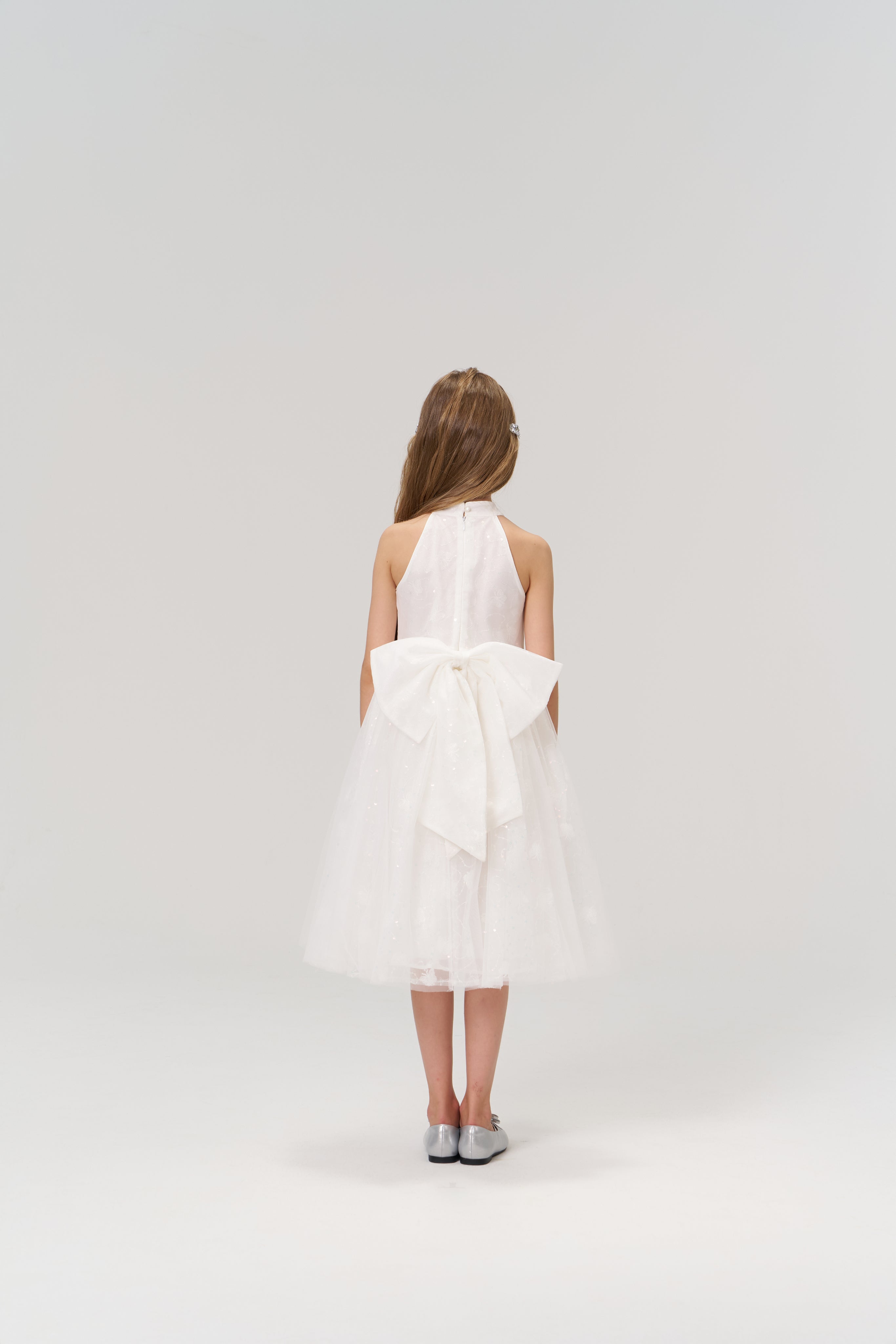 A young girl with long hair faces away, wearing the KIKI and JOJO Butterfly Whispers Dress—a white halter dress with a large back bow—paired with silver shoes, set against a plain light background.