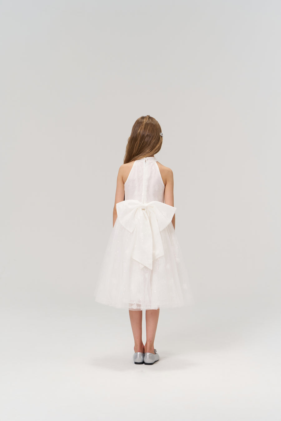 A young girl with long hair faces away, wearing the KIKI and JOJO Butterfly Whispers Dress—a white halter dress with a large back bow—paired with silver shoes, set against a plain light background.
