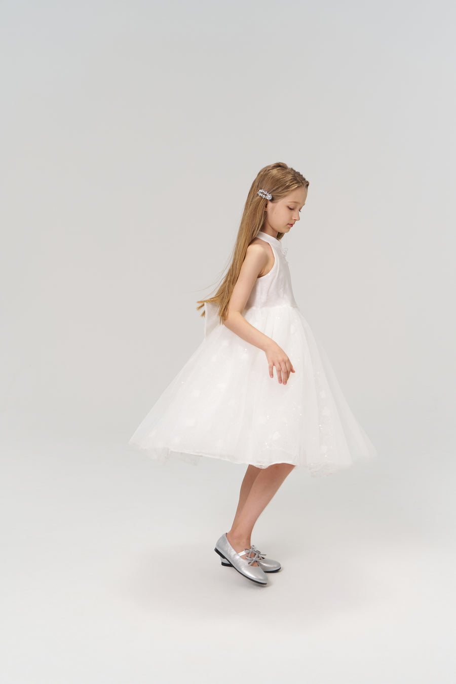 A young girl wearing the KIKI and JOJO Butterfly Whispers Dress, featuring a white butterfly halter top and layered tulle skirt, pairs it with silver shoes while standing sideways against a plain light background.