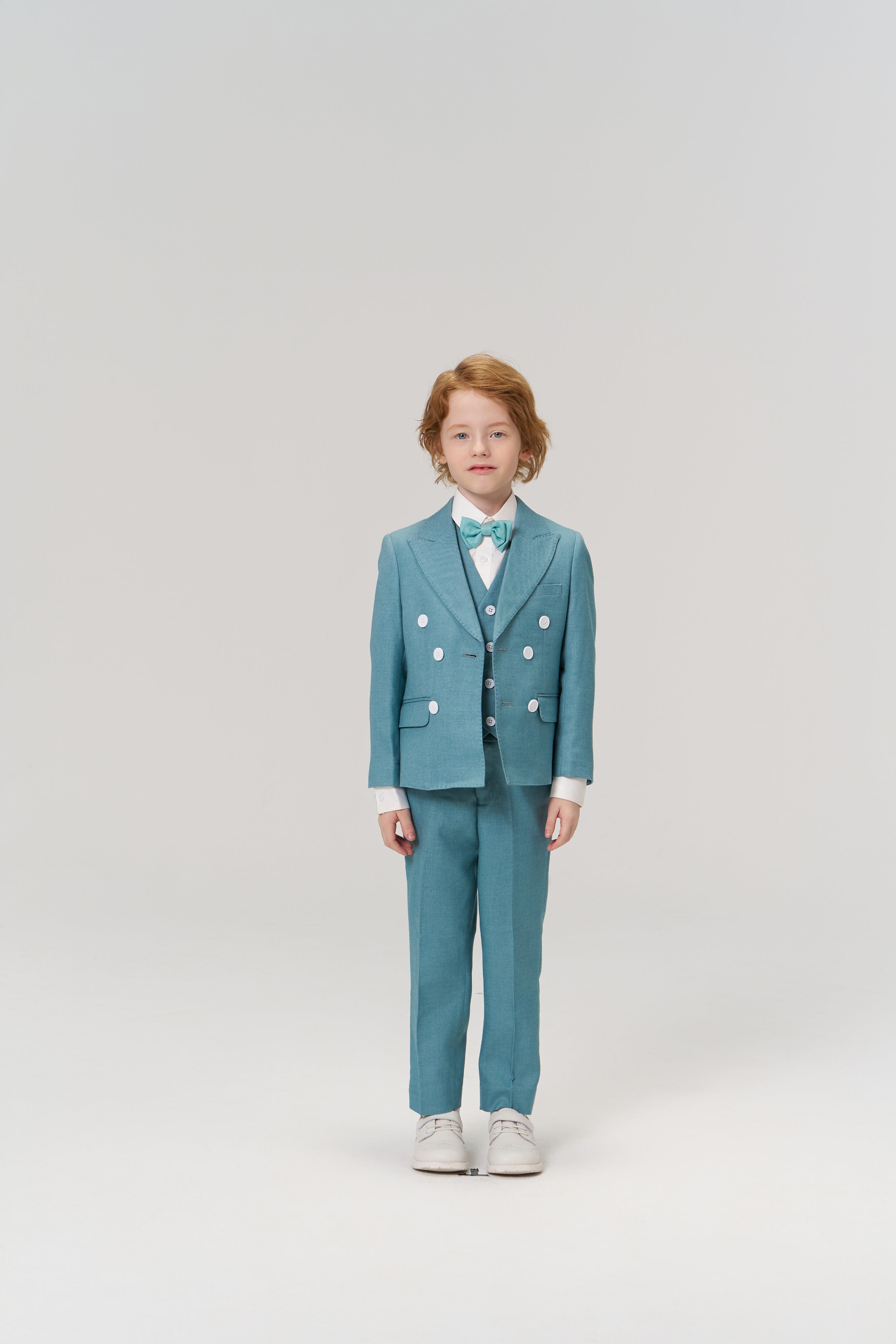 A young child with light skin and red hair faces forward, wearing the KIKI and JOJO Celeste Whimsy Suit Pants with an adjustable waistband, a turquoise suit, white shirt, bow tie, and white shoes against a plain light background.
