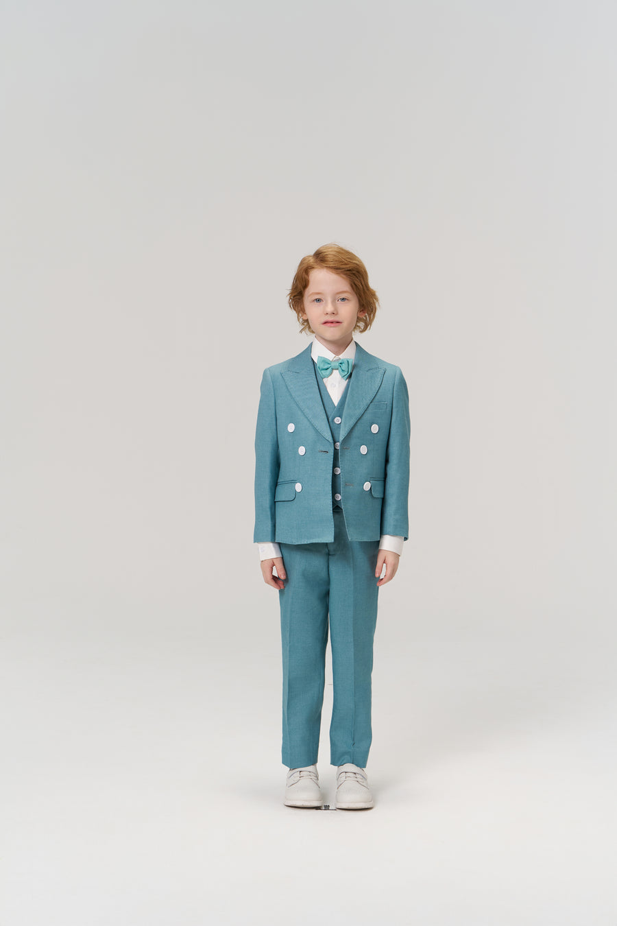 A young child with light skin and red hair faces forward, wearing the KIKI and JOJO Celeste Whimsy Suit Pants with an adjustable waistband, a turquoise suit, white shirt, bow tie, and white shoes against a plain light background.