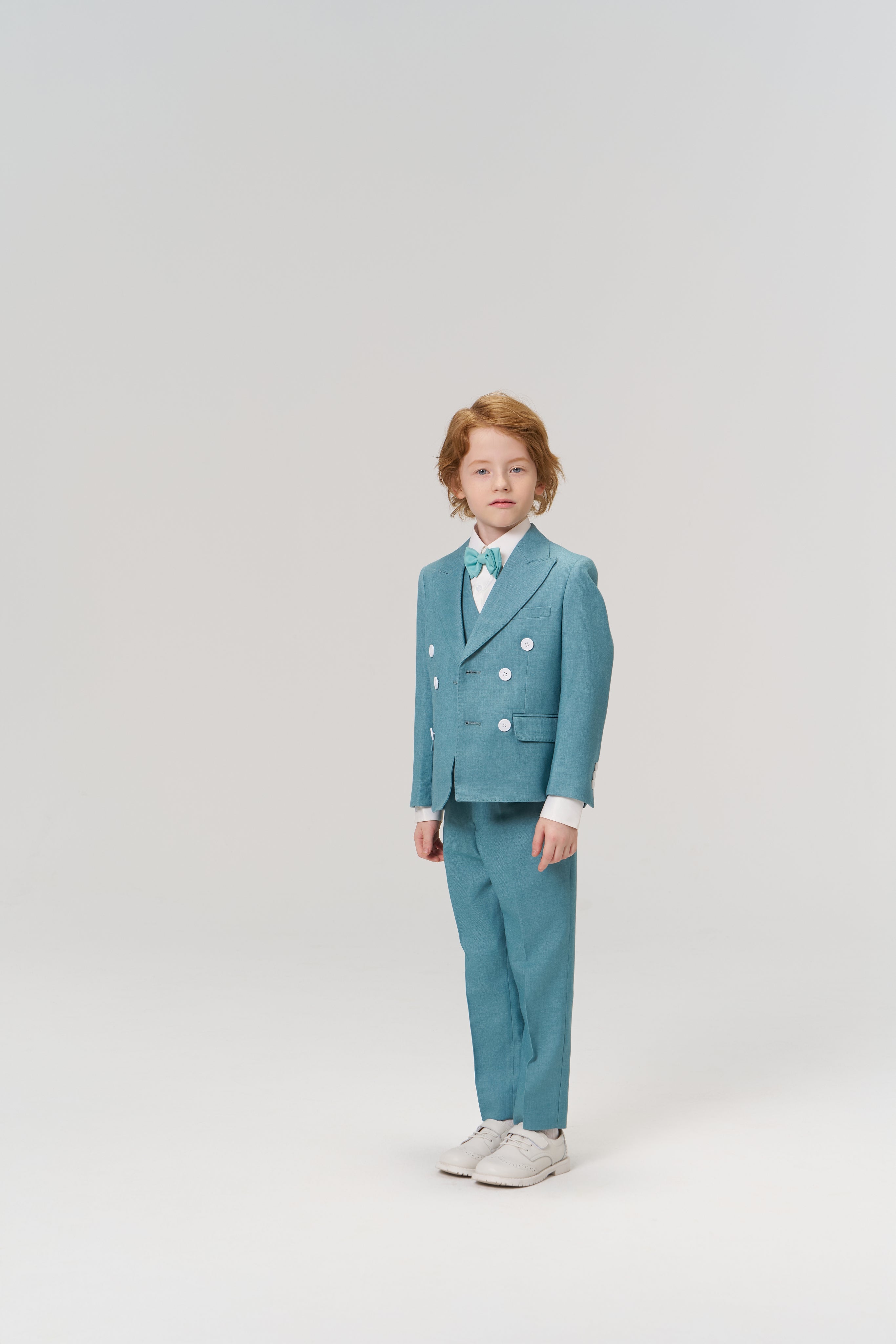 A young child stands against a plain background, wearing KIKI and JOJO’s Celeste Whimsy Suit Pants with an adjustable waistband, paired with a turquoise suit jacket, bow tie, and white sneakers.