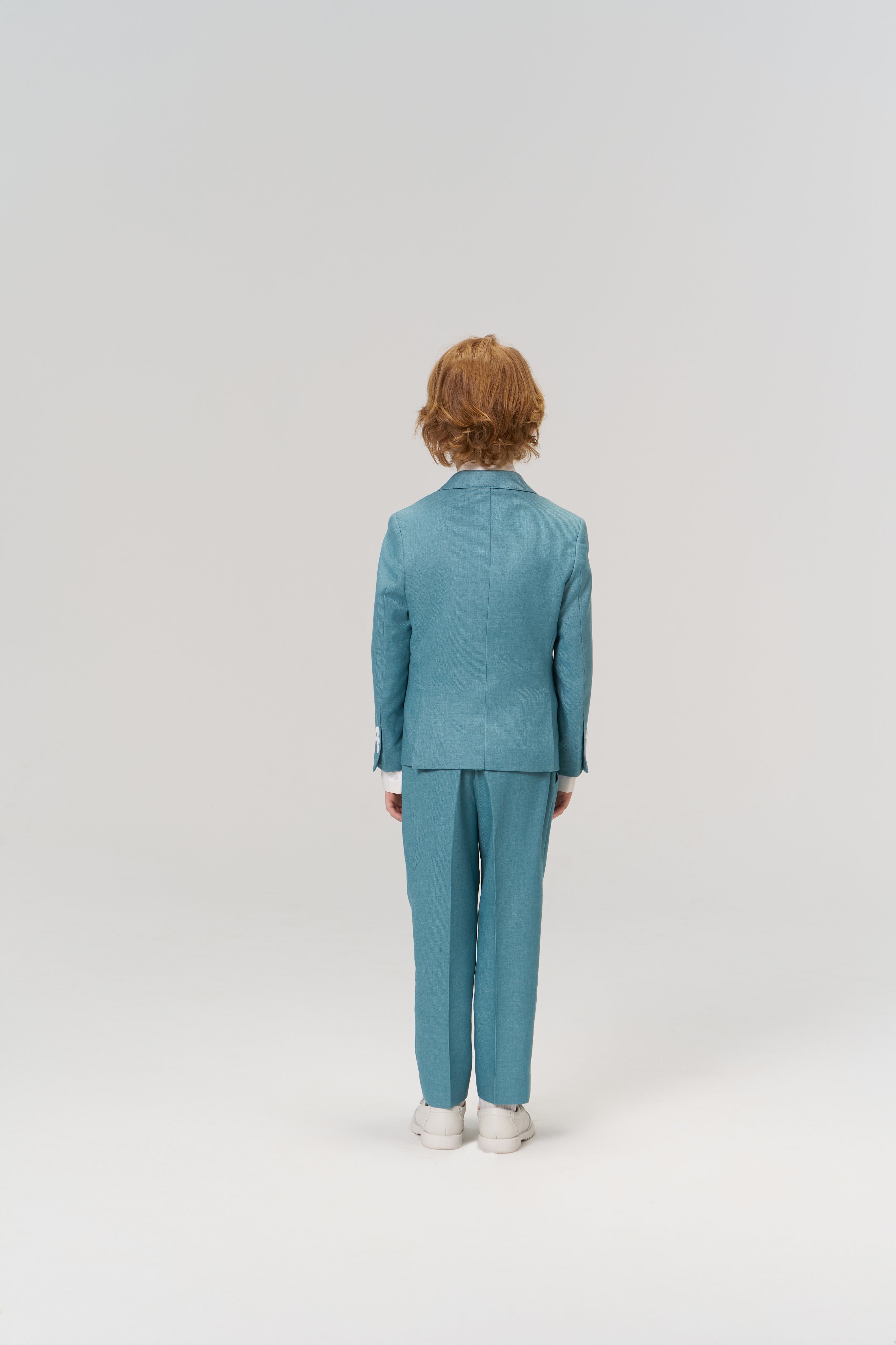 A child with shoulder-length hair, dressed in KIKI and JOJO's Celeste Whimsy Suit Pants with an adjustable waistband and white shoes, stands facing away from the camera against a plain light background.
