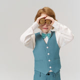 A young boy wears KIKI and JOJO’s Celeste Whimsy Suit Pants with an adjustable waistband and bow tie, playfully holding two cookies to his eyes like binoculars against a plain light background.