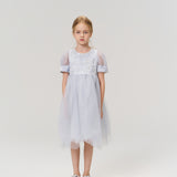 A young girl with light brown hair wears the KIKI and JOJO Mist Whisper Dress, featuring hand-embroidered florals, and silver shoes as she stands against a plain light background, looking directly at the camera.