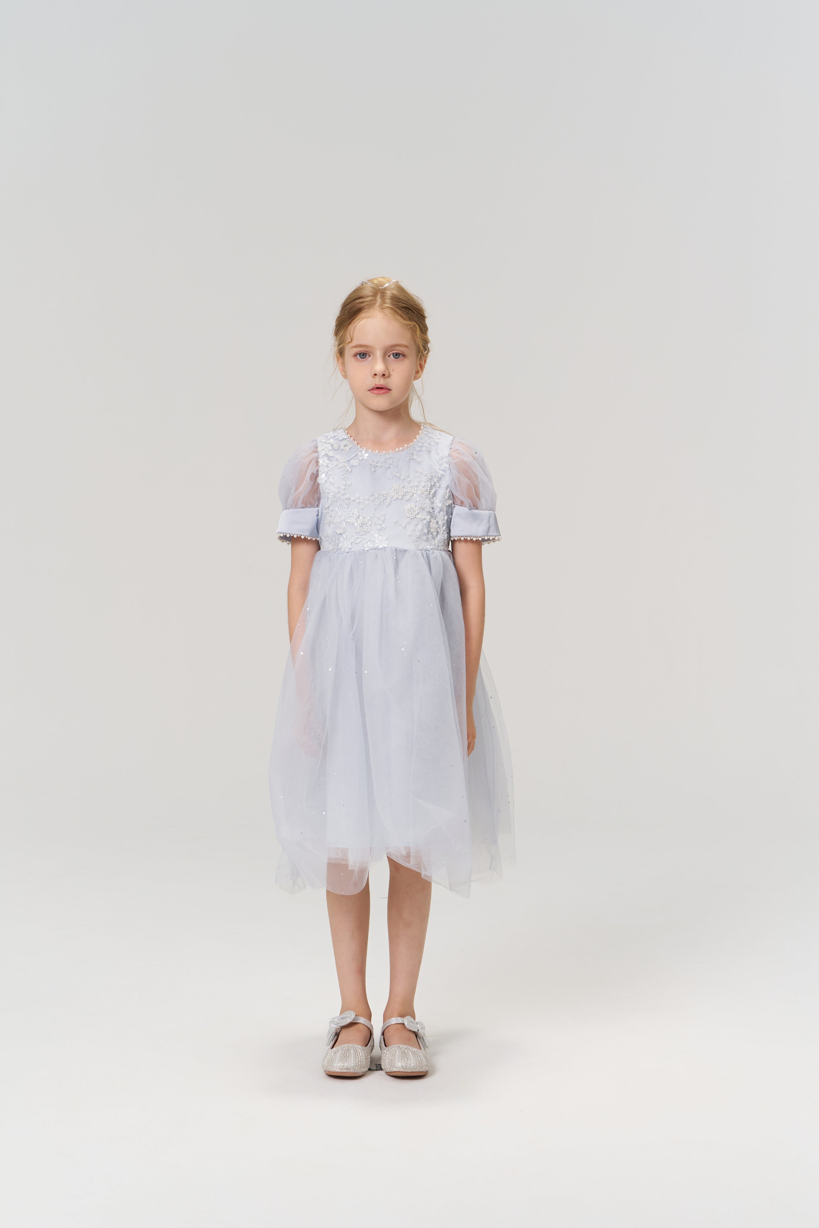 A young girl with light brown hair wears the KIKI and JOJO Mist Whisper Dress, featuring hand-embroidered florals, and silver shoes as she stands against a plain light background, looking directly at the camera.