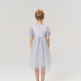 A young girl with blonde hair in a bun stands facing away against a plain light background, wearing the KIKI and JOJO Mist Whisper Dress in light blue and silver shoes.