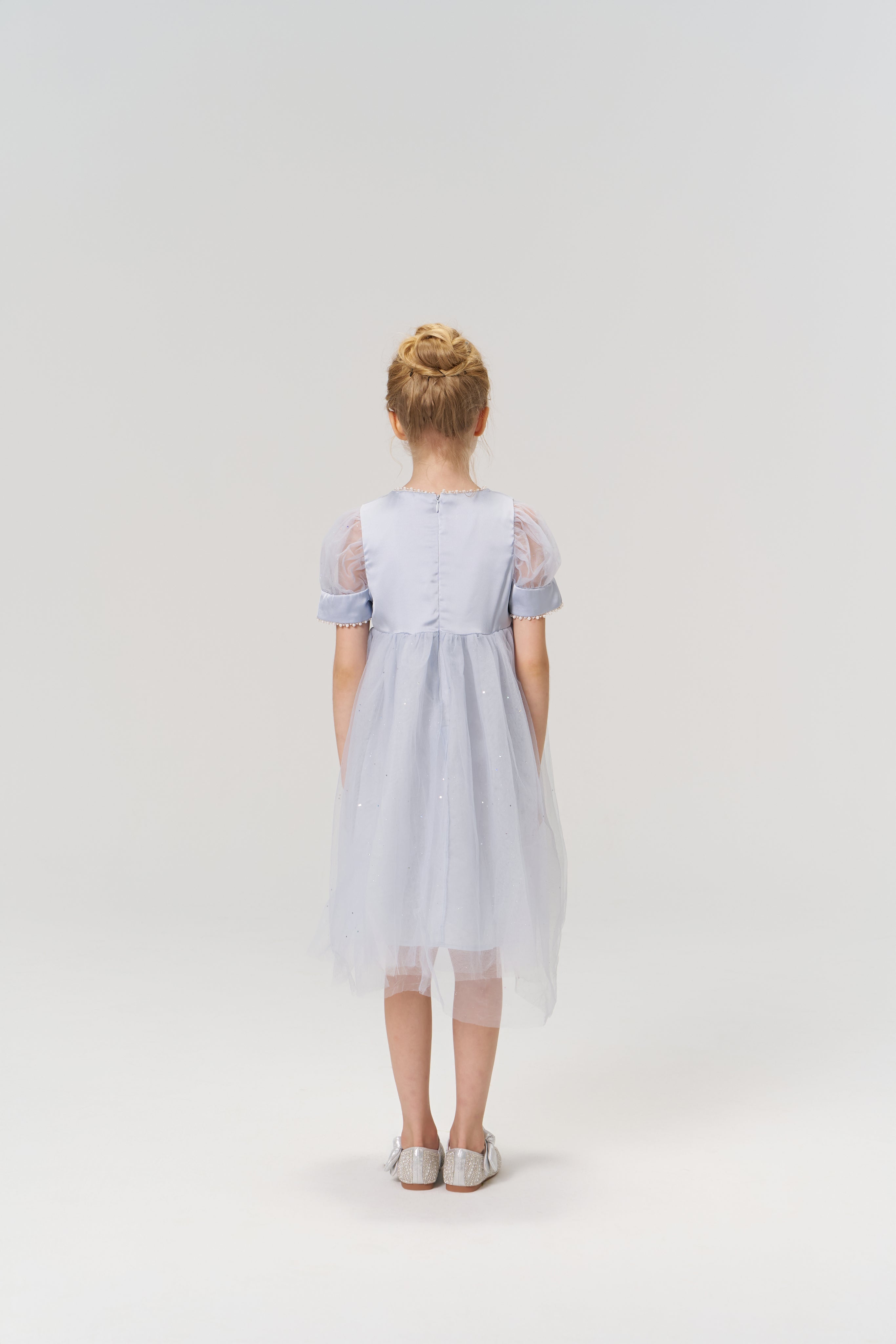 A young girl with blonde hair in a bun stands facing away against a plain light background, wearing the KIKI and JOJO Mist Whisper Dress in light blue and silver shoes.