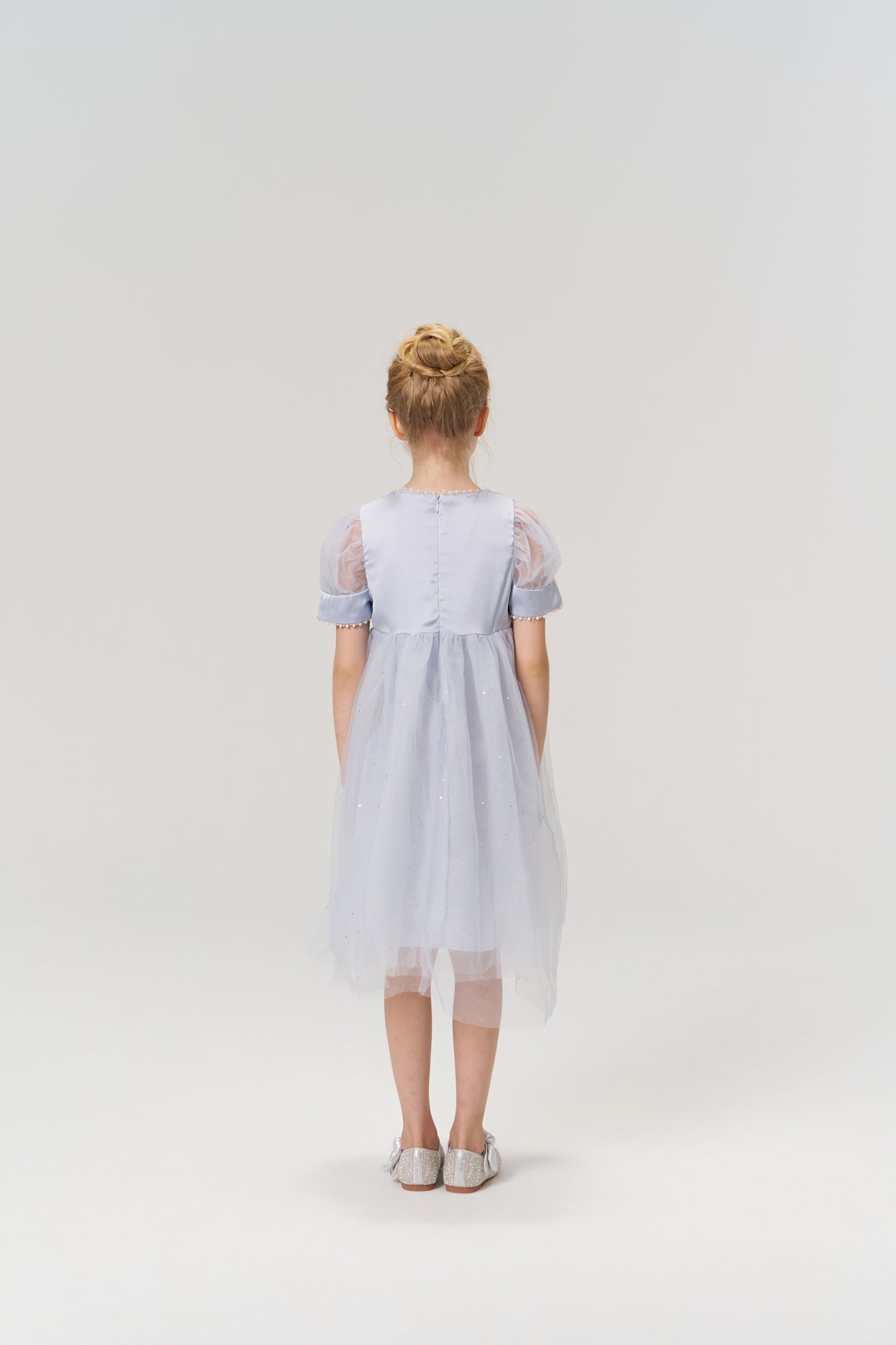 A young girl with blonde hair in a bun stands facing away against a plain light background, wearing the KIKI and JOJO Mist Whisper Dress in light blue and silver shoes.