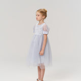A young girl with blonde hair in a bun stands sideways against a plain light background, wearing the Mist Whisper Dress by KIKI and JOJO—a light blue A-line dress with puffed sleeves.