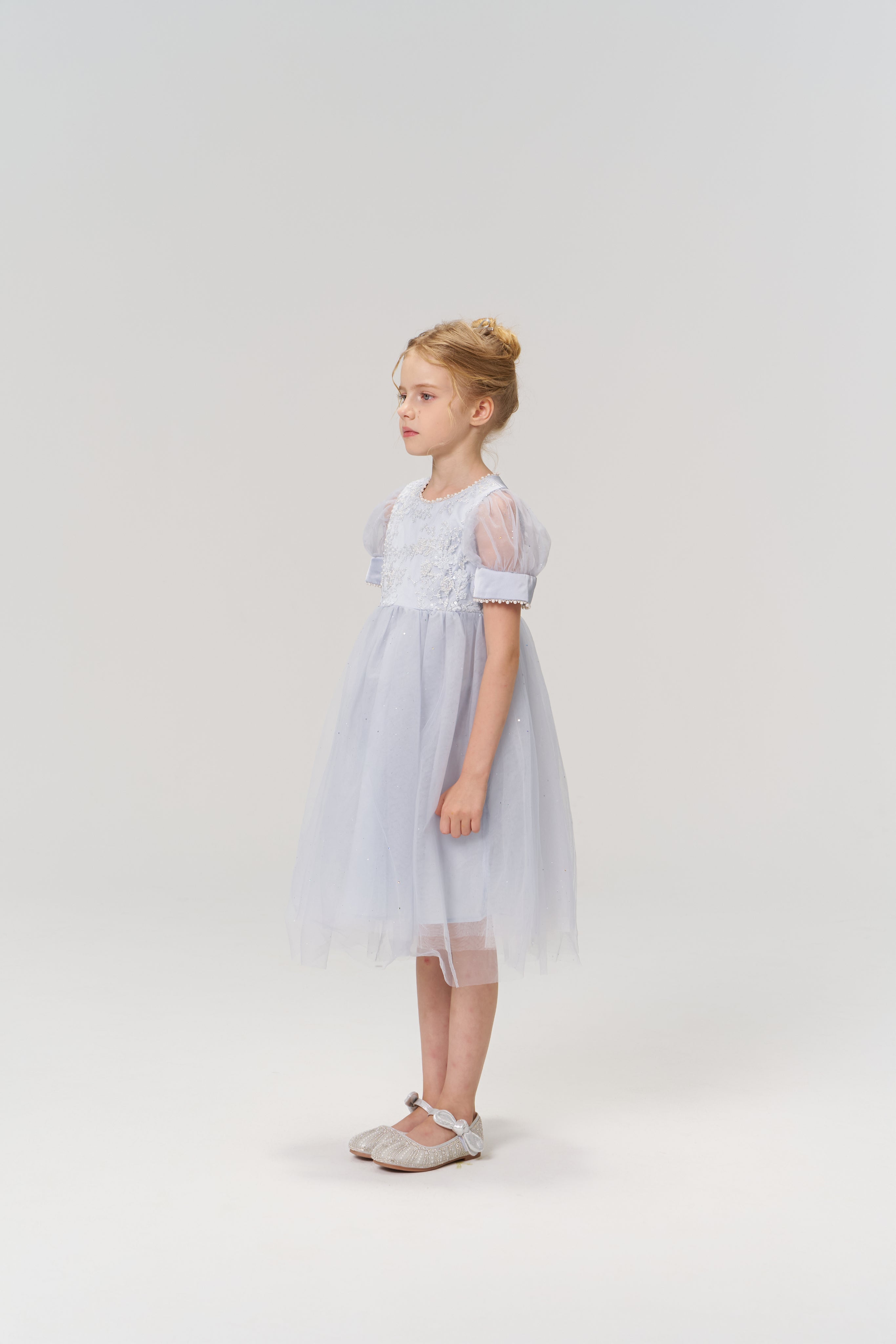 A young girl with blonde hair in a bun stands sideways against a plain light background, wearing the Mist Whisper Dress by KIKI and JOJO—a light blue A-line dress with puffed sleeves.