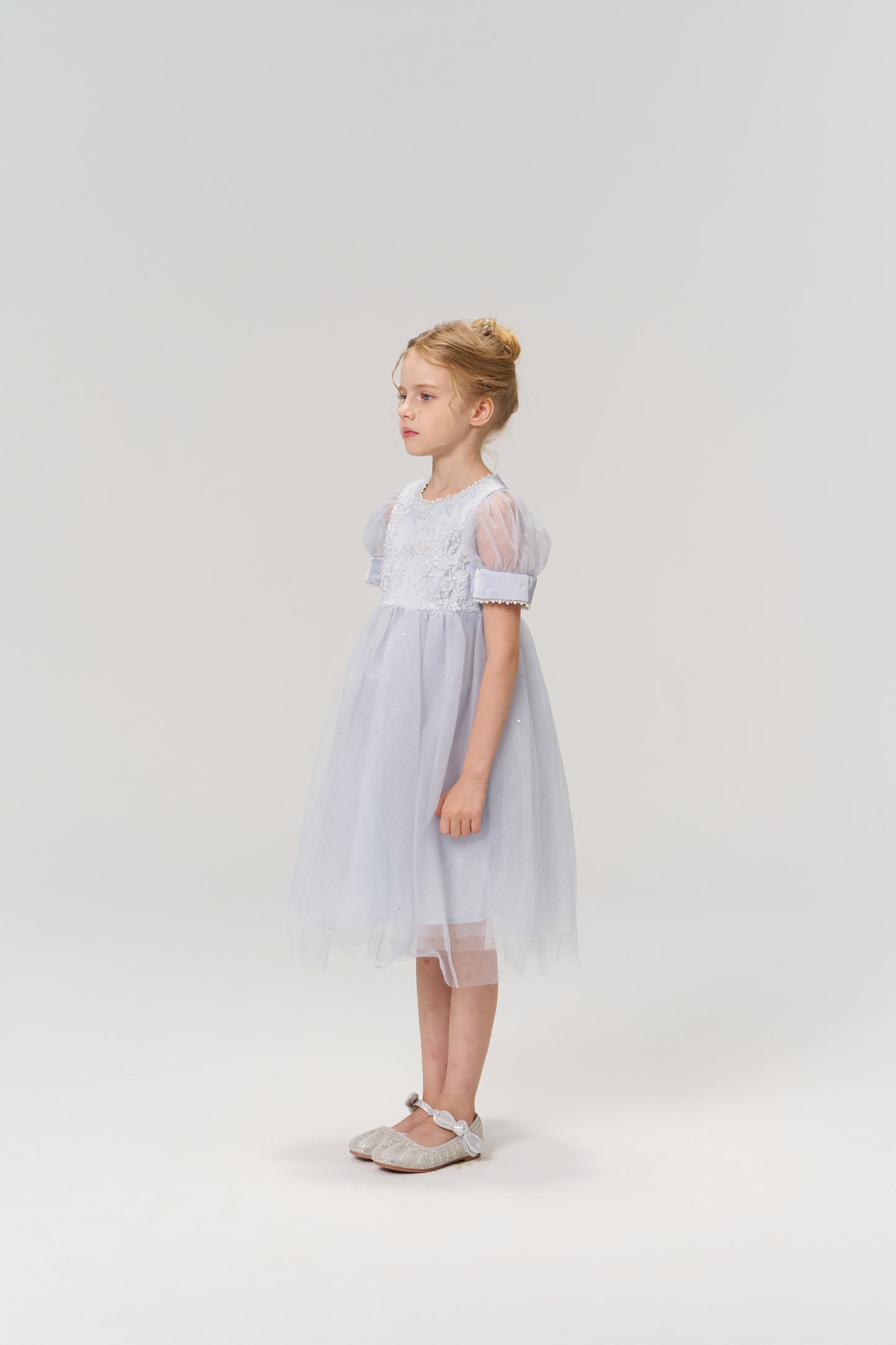 A young girl with blonde hair in a bun stands sideways against a plain light background, wearing the Mist Whisper Dress by KIKI and JOJO—a light blue A-line dress with puffed sleeves.