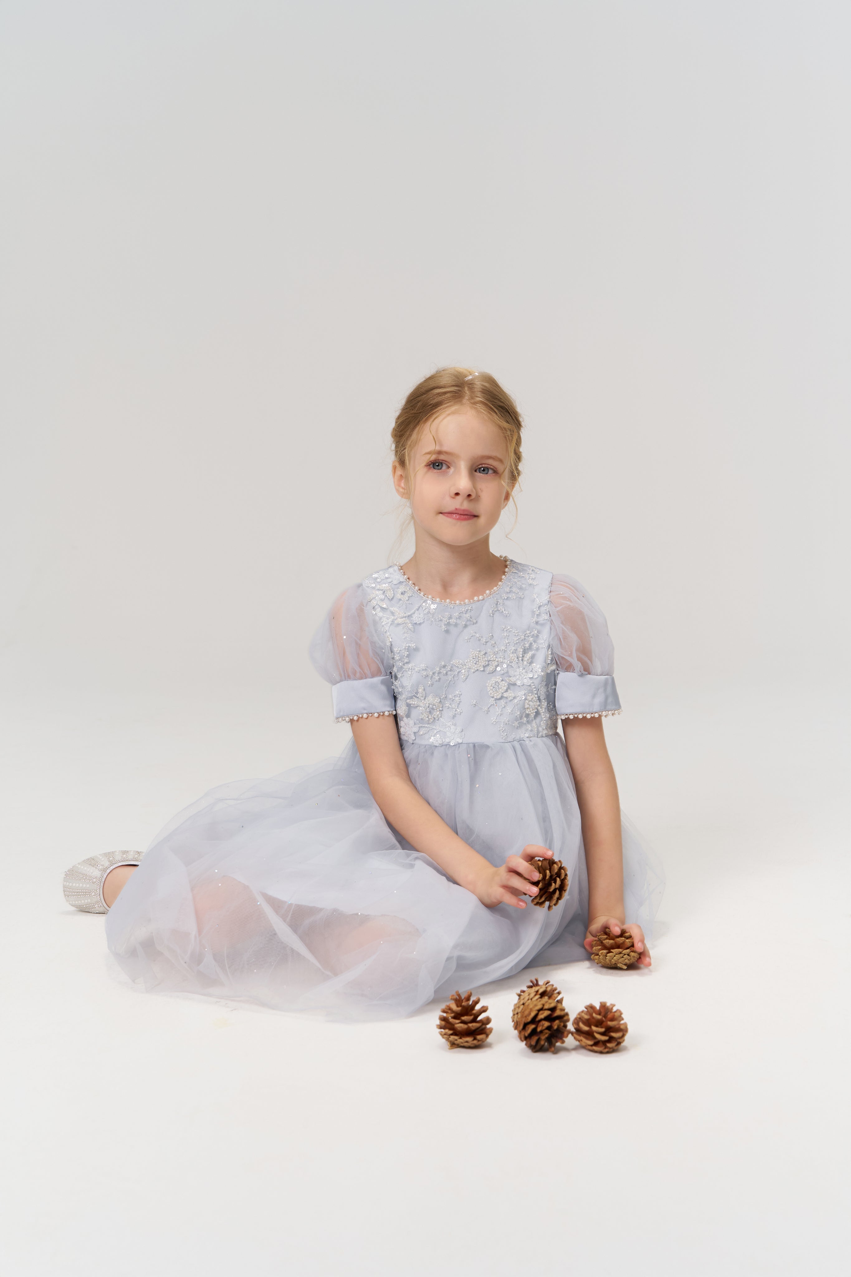Young girl in a light blue dress sits on the floor holding pine cones, with more pine cones placed in front of her, against a plain white background.