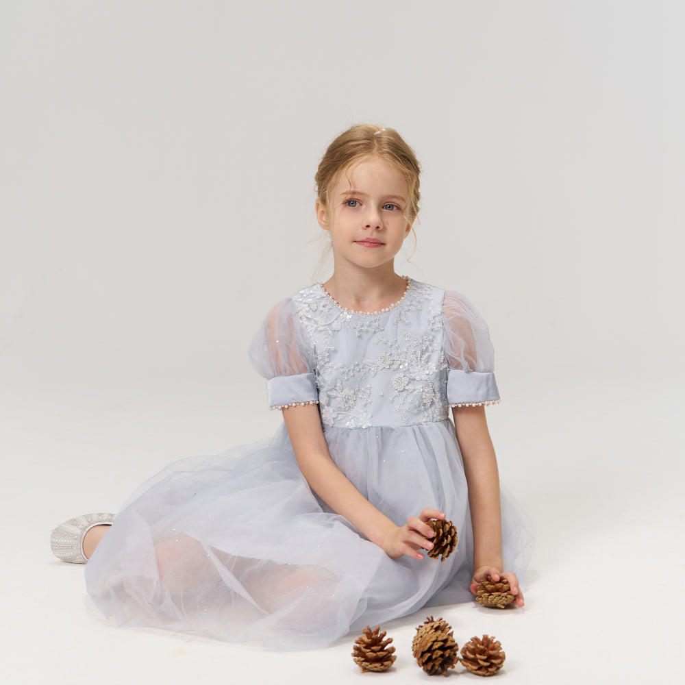 A young girl in a light blue dress sits on the floor holding pine cones, with more pine cones placed in front of her on a white background.