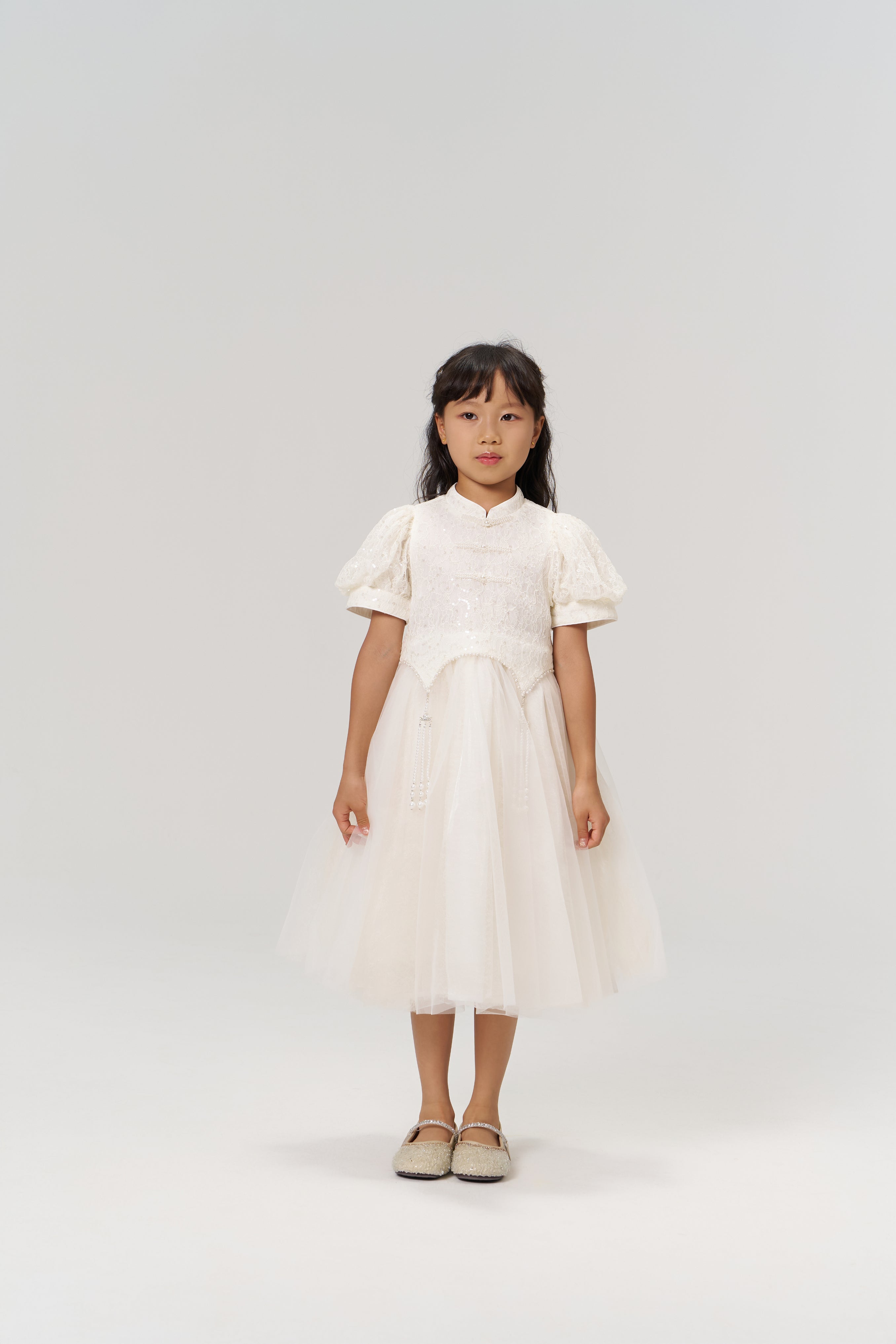 A young girl faces the camera wearing the KIKI and JOJO Orchid Whisper Dress, a modern vintage white dress with sequin embroidered lace, paired with beige shoes against a plain light background.