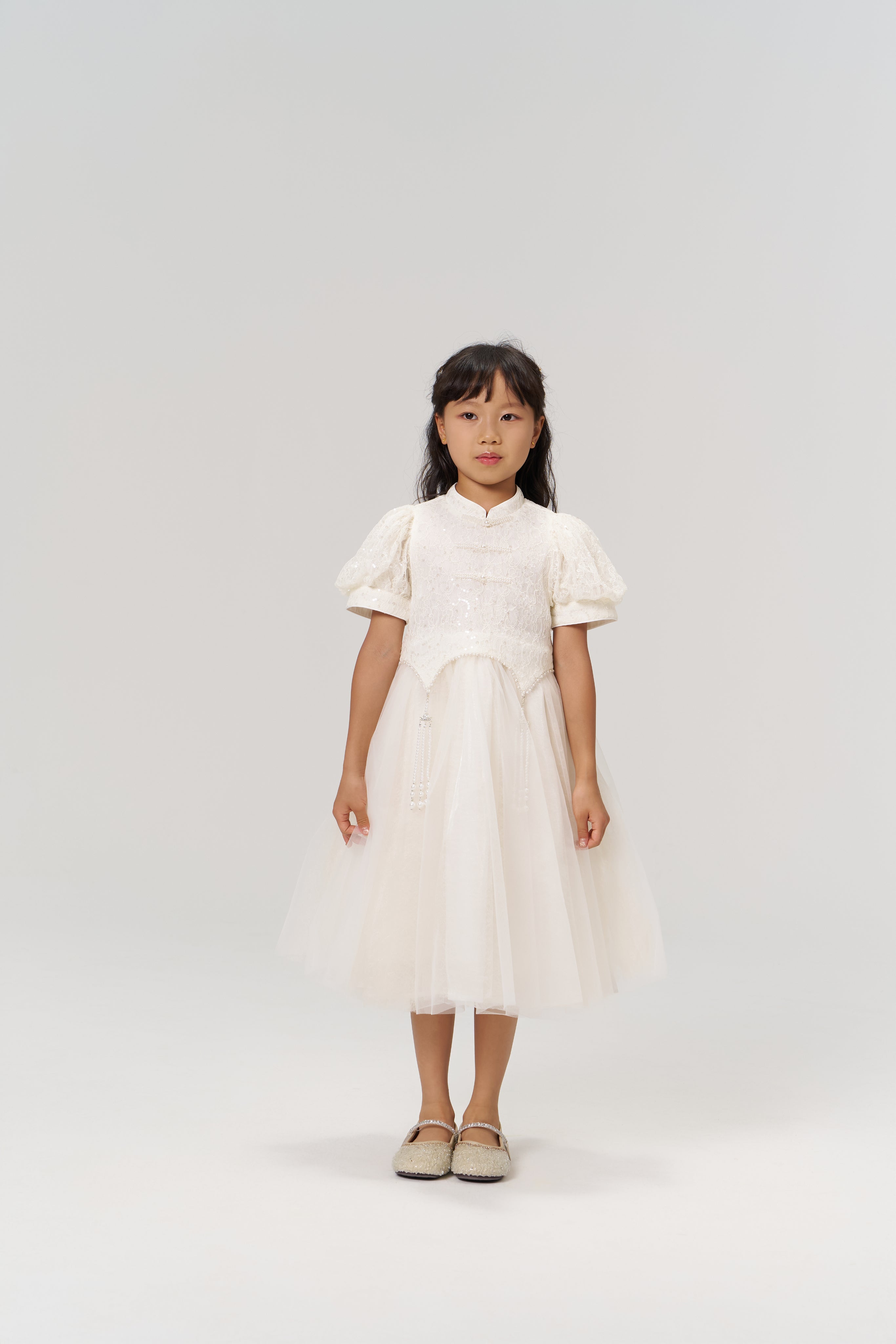 A young girl faces the camera wearing the KIKI and JOJO Orchid Whisper Dress, a modern vintage white dress with sequin embroidered lace, paired with beige shoes against a plain light background.