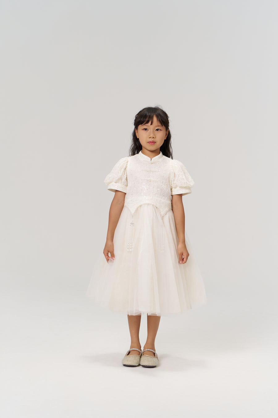 A young girl faces the camera wearing the KIKI and JOJO Orchid Whisper Dress, a modern vintage white dress with sequin embroidered lace, paired with beige shoes against a plain light background.