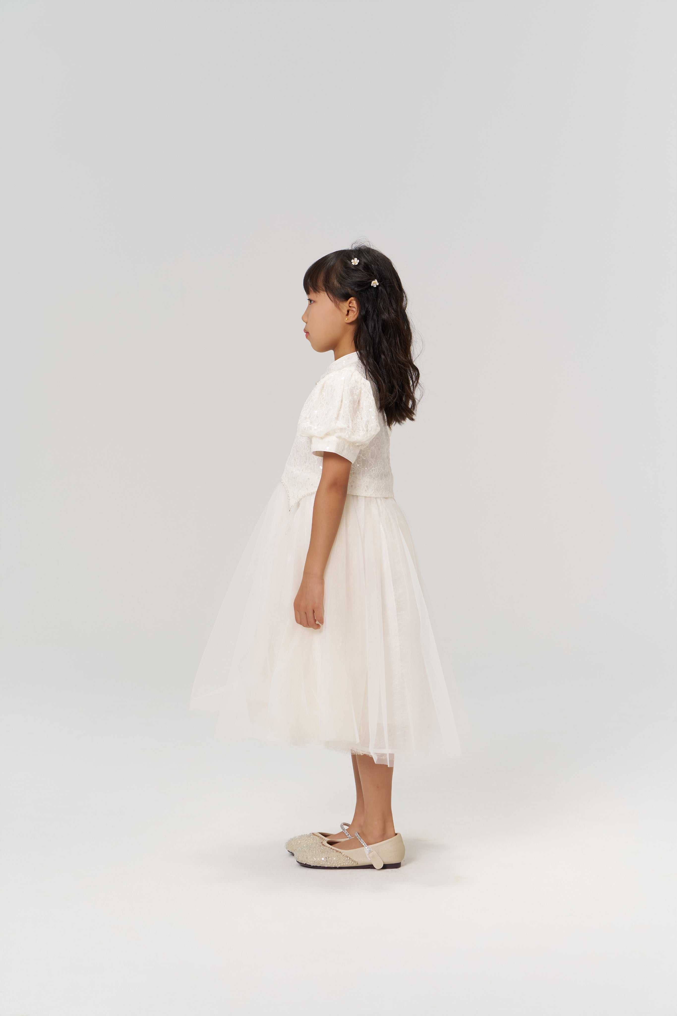 A young girl in profile wears the Orchid Whisper Dress by KIKI and JOJO—featuring a modern vintage style with a tulle skirt—and beige shoes, set against a plain light background.