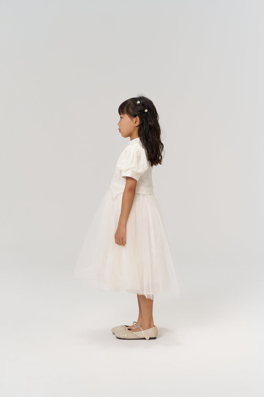 A young girl in profile wears the Orchid Whisper Dress by KIKI and JOJO—featuring a modern vintage style with a tulle skirt—and beige shoes, set against a plain light background.