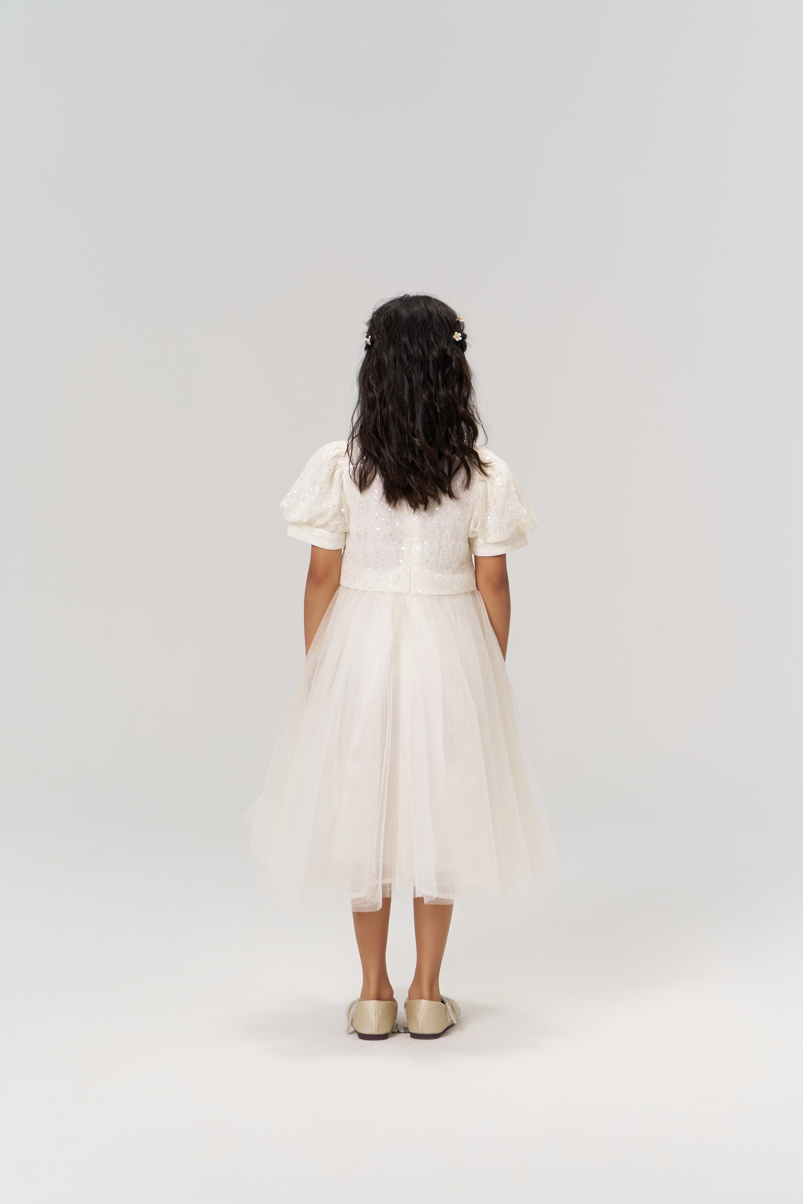 A young girl wears the KIKI and JOJO Orchid Whisper Dress, featuring sequin-embroidered lace and matching shoes, standing with her back to the camera against a plain light background.