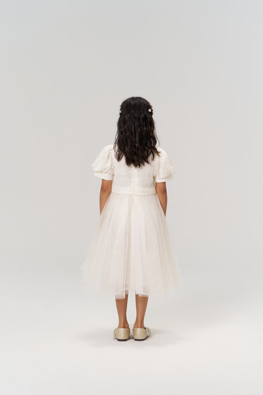 A young girl wears the KIKI and JOJO Orchid Whisper Dress, featuring sequin-embroidered lace and matching shoes, standing with her back to the camera against a plain light background.