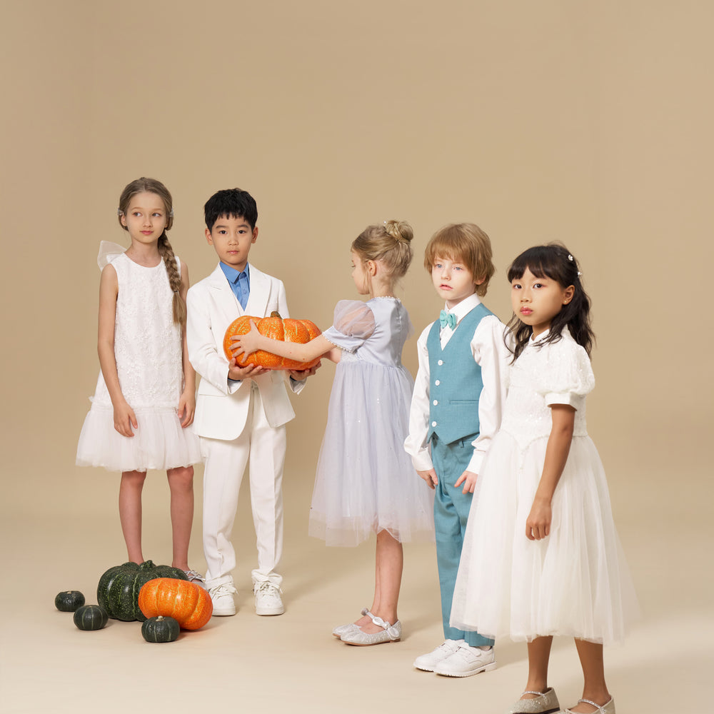Five children in formal attire stand indoors with pumpkins on the floor; one child holds two orange pumpkins. Background is plain beige.