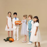 Five children in formal attire stand indoors with pumpkins on the floor; one child holds two orange pumpkins. Background is plain beige.