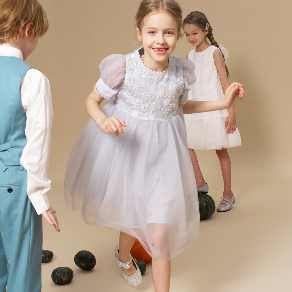 Three children play together indoors; one girl in a light blue dress is in motion, while another girl in white and a boy in a blue vest look on. Several small gourds are on the floor.