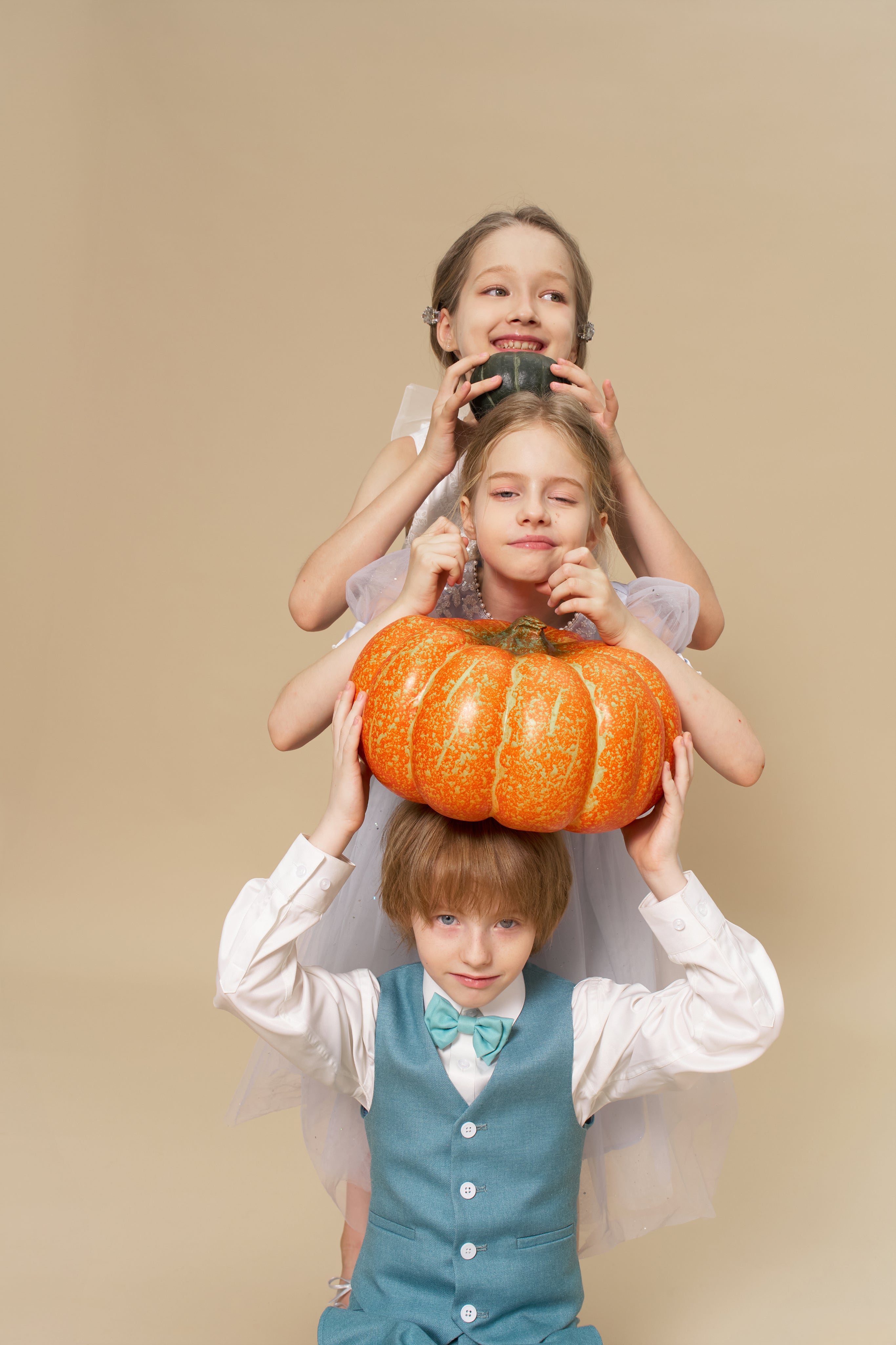 Three children pose in a stack against a beige background; the middle, wearing KIKI and JOJO Celeste Whimsy Suit Pants with an adjustable waistband, balances a pot on their head while the others hold another pot and a pumpkin.