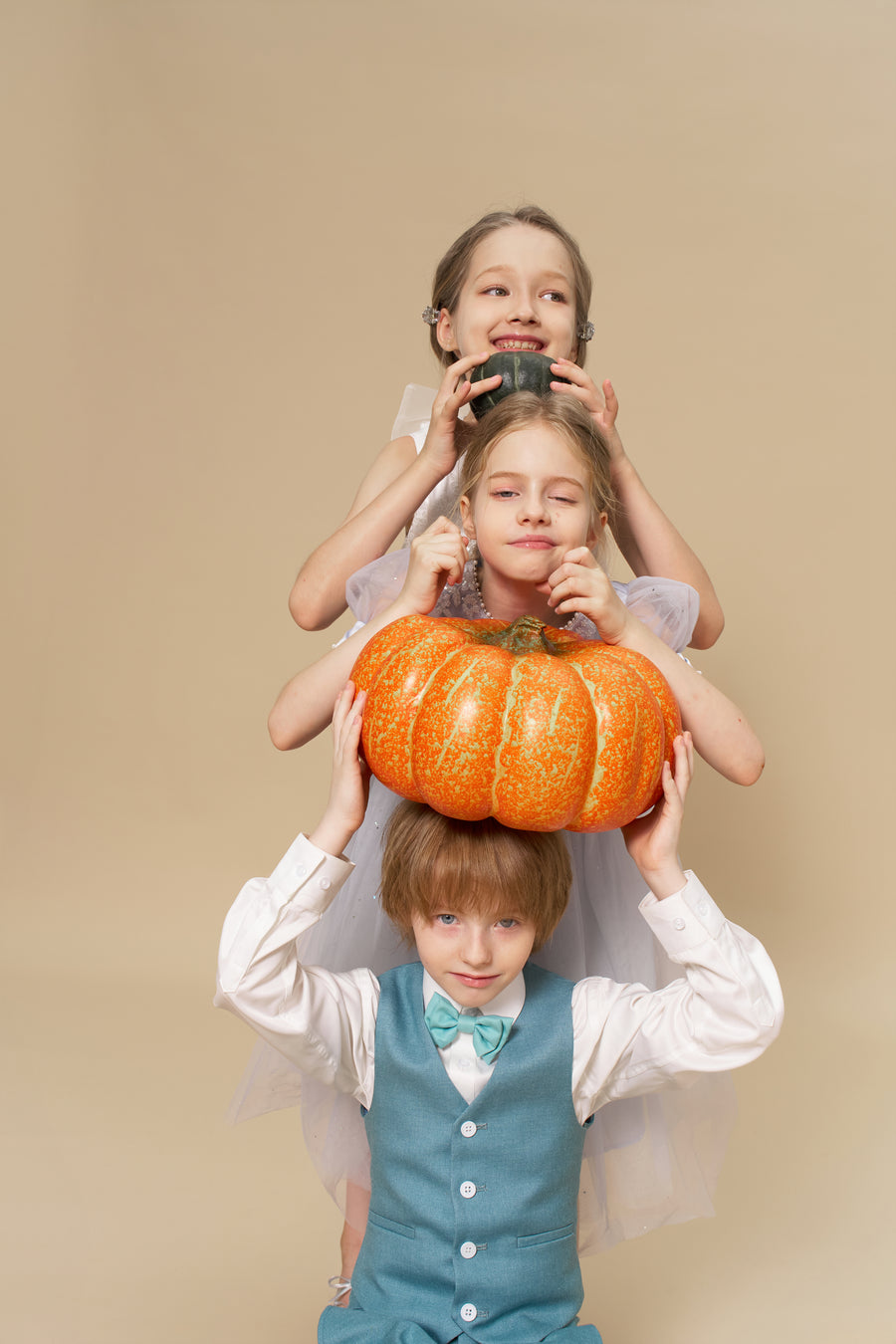Three children pose in a stack against a beige background; the middle, wearing KIKI and JOJO Celeste Whimsy Suit Pants with an adjustable waistband, balances a pot on their head while the others hold another pot and a pumpkin.
