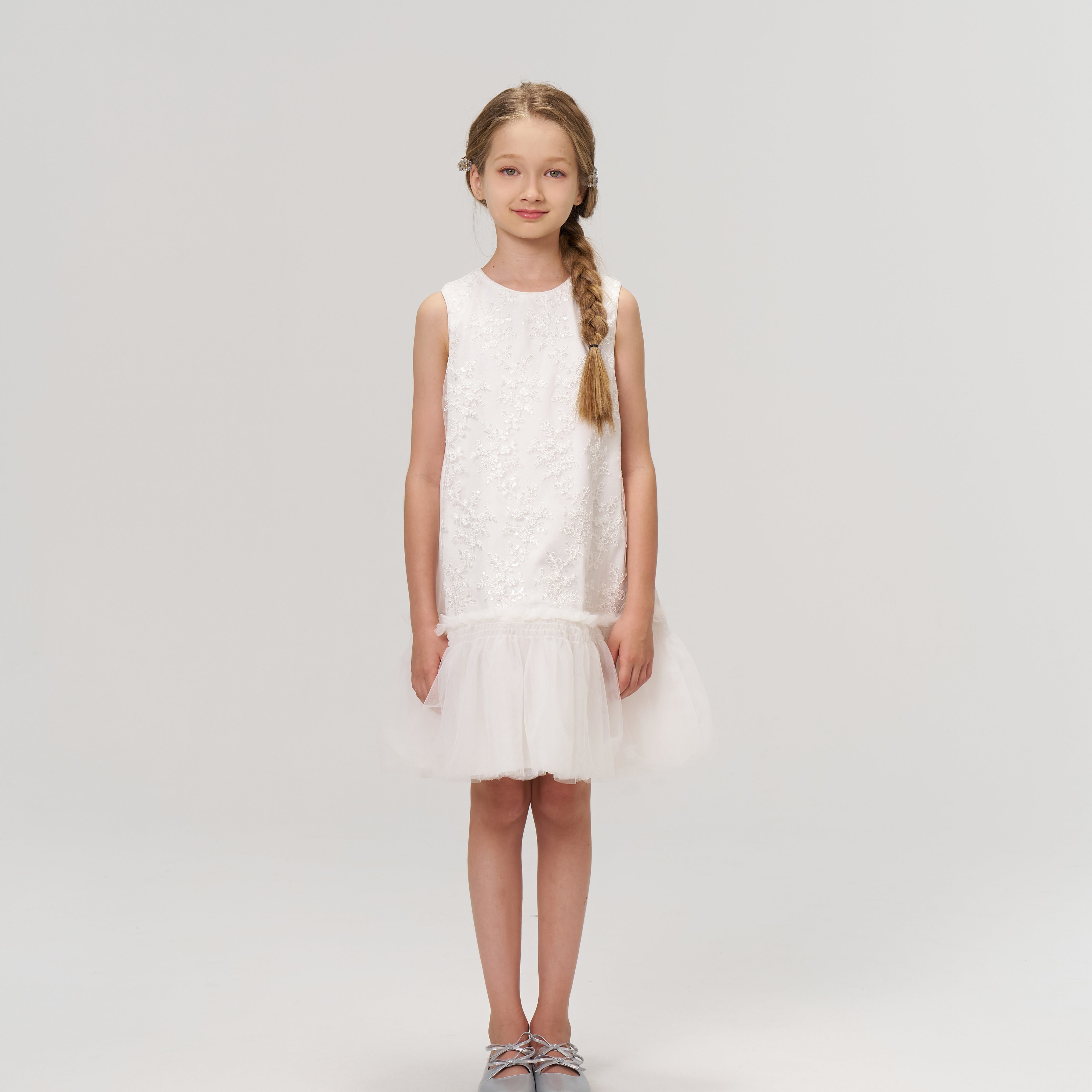 A young girl with a braid faces forward, wearing the KIKI and JOJO Twinkle Whisper Dress—a sleeveless white A-line dress with a tulle hem, embroidered lace, and silver shoes—set against a plain light background.