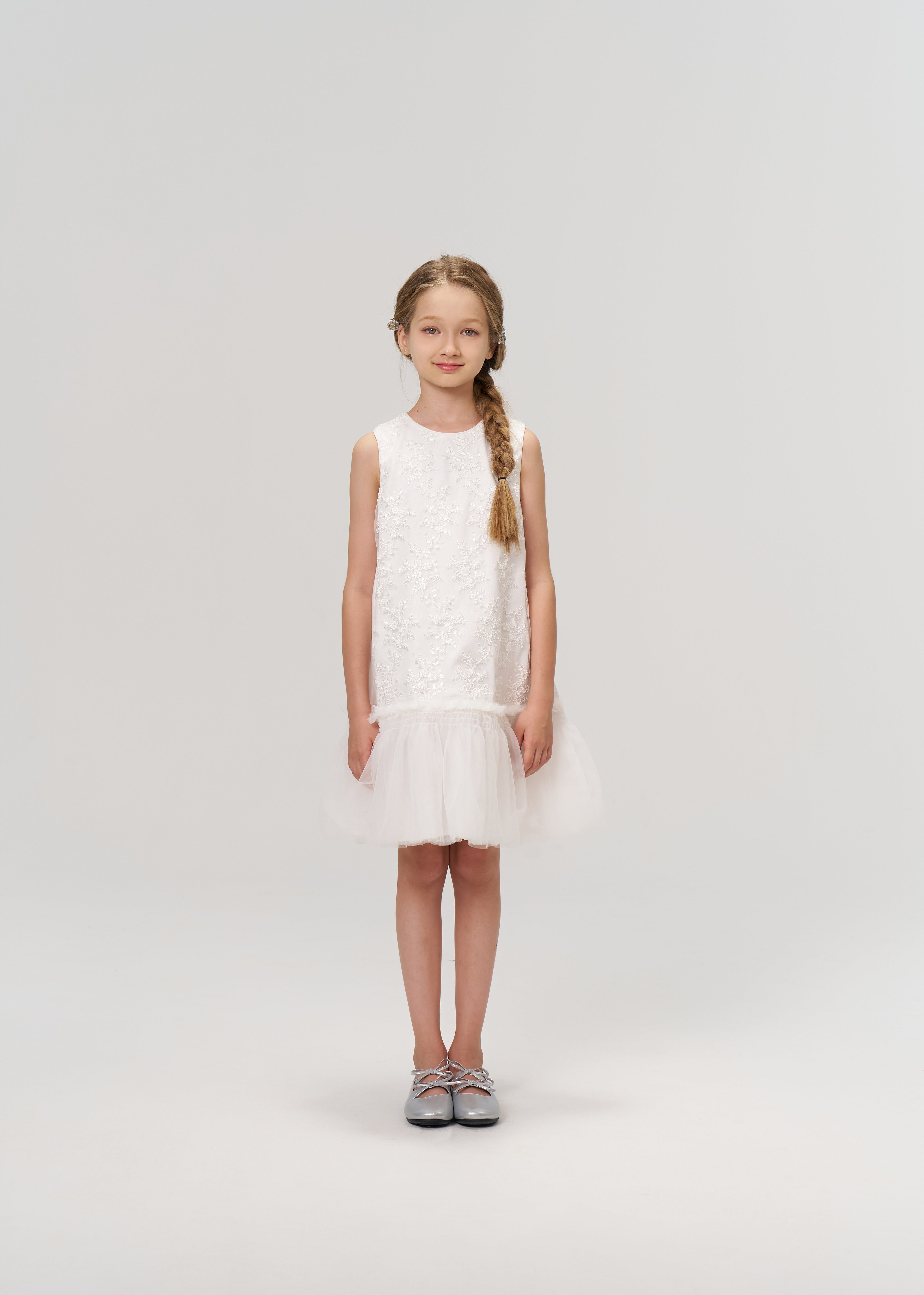A young girl with a braid faces forward, wearing the KIKI and JOJO Twinkle Whisper Dress—a sleeveless white A-line dress with a tulle hem, embroidered lace, and silver shoes—set against a plain light background.