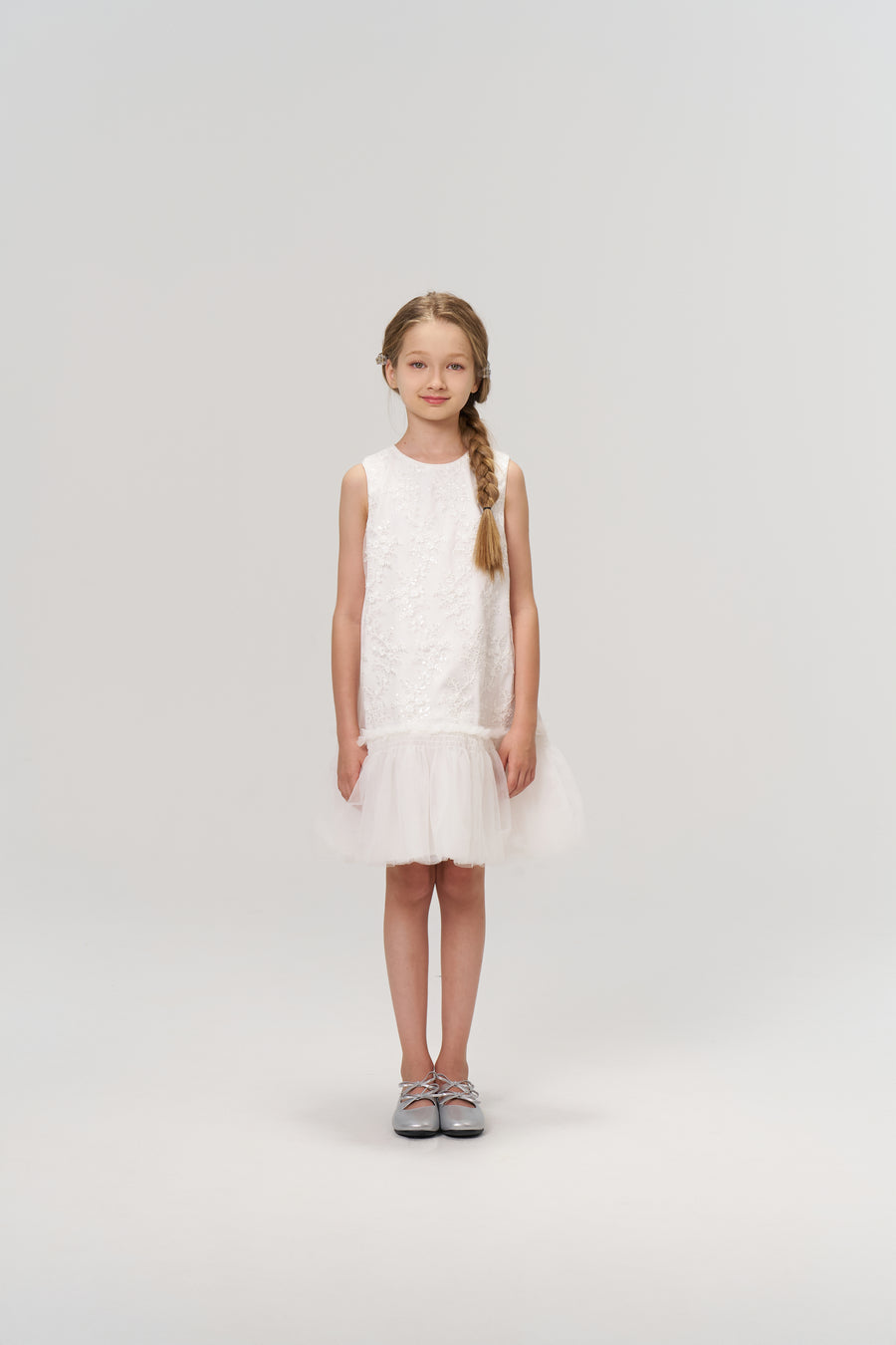 A young girl with a braid faces forward, wearing the KIKI and JOJO Twinkle Whisper Dress—a sleeveless white A-line dress with a tulle hem, embroidered lace, and silver shoes—set against a plain light background.
