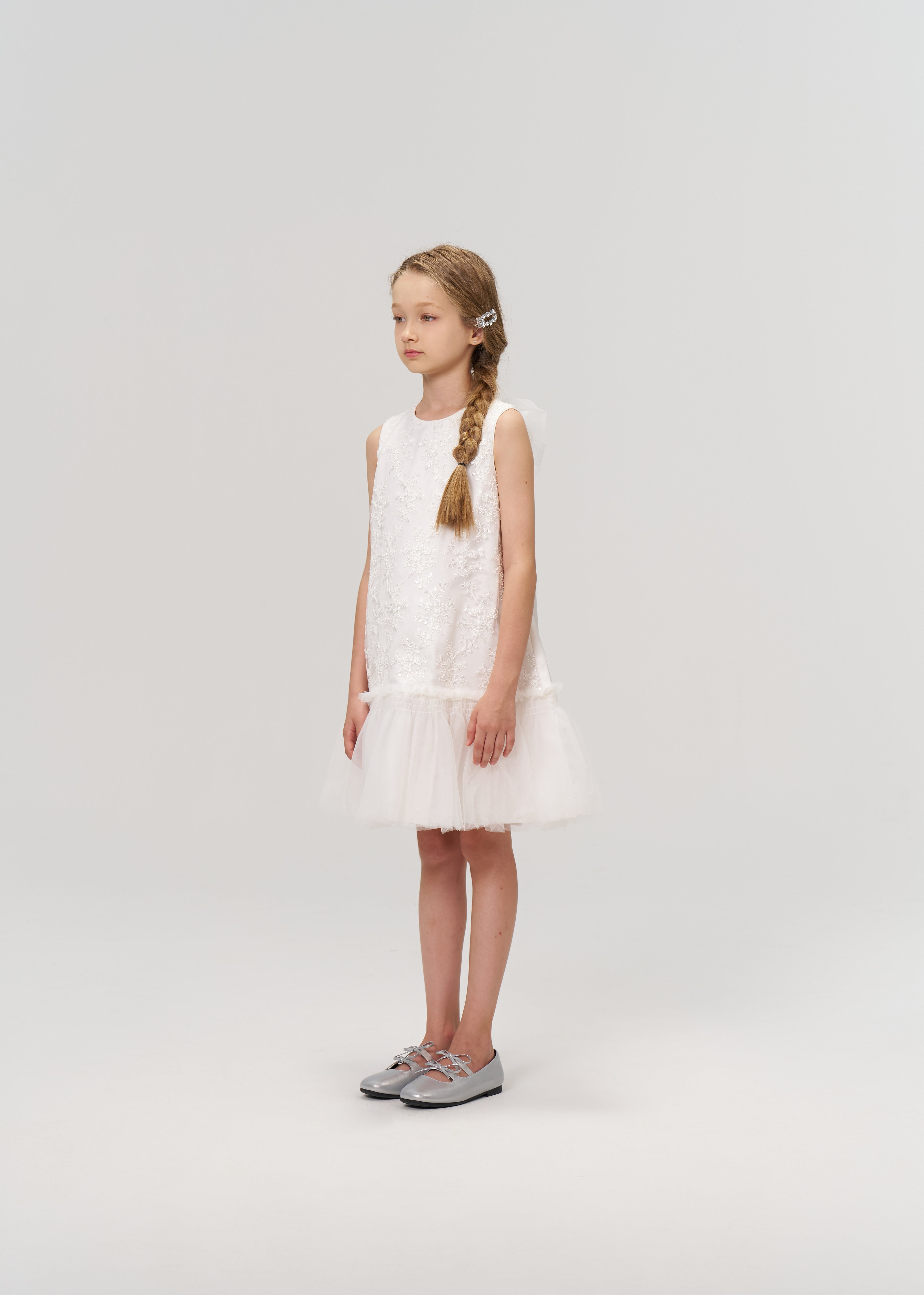 A young girl with a side braid stands in profile wearing the KIKI and JOJO Twinkle Whisper Dress, a sleeveless white A-line style with an embroidered lace tulle hem, paired with silver shoes against a plain light background.