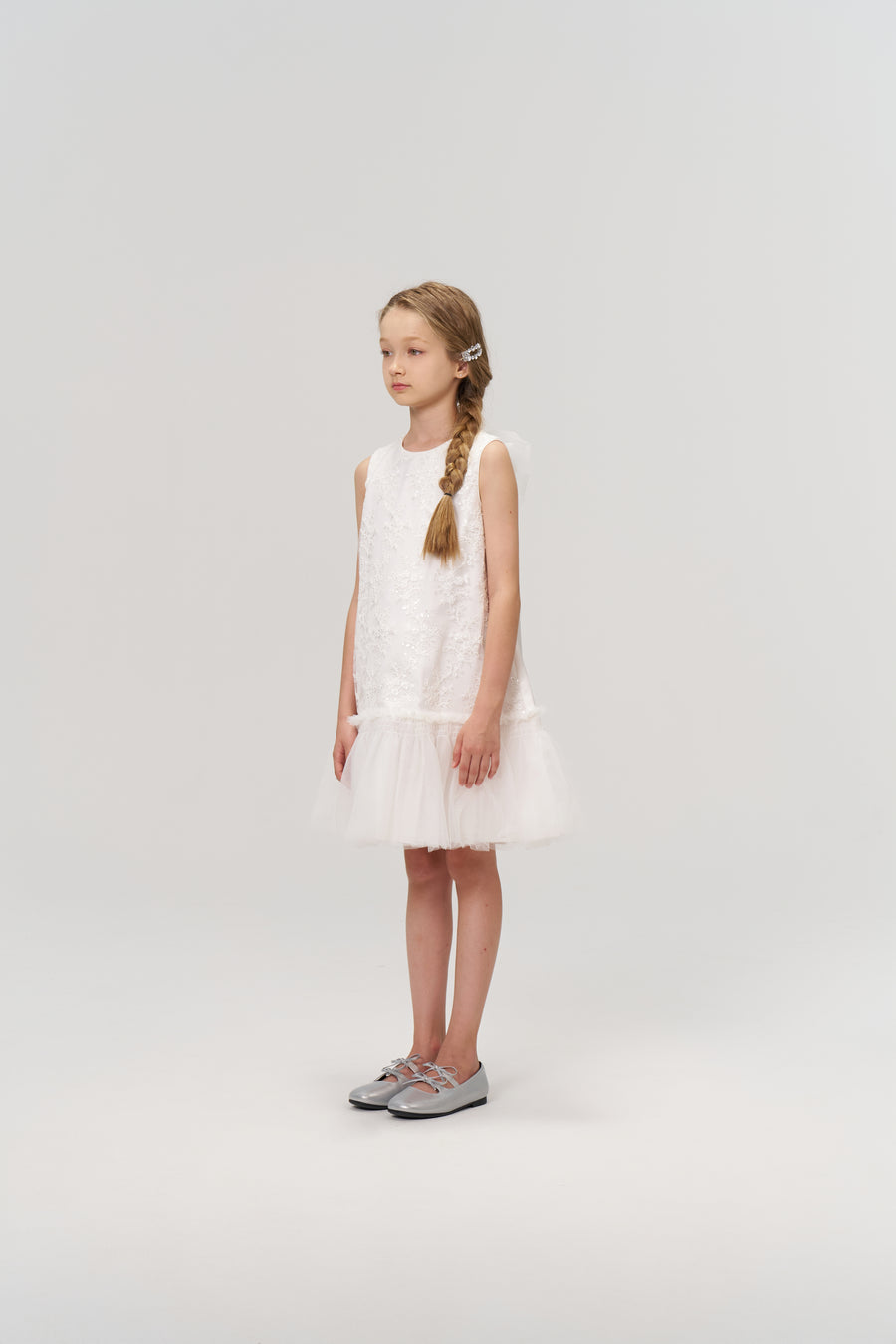 A young girl with a side braid stands in profile wearing the KIKI and JOJO Twinkle Whisper Dress, a sleeveless white A-line style with an embroidered lace tulle hem, paired with silver shoes against a plain light background.