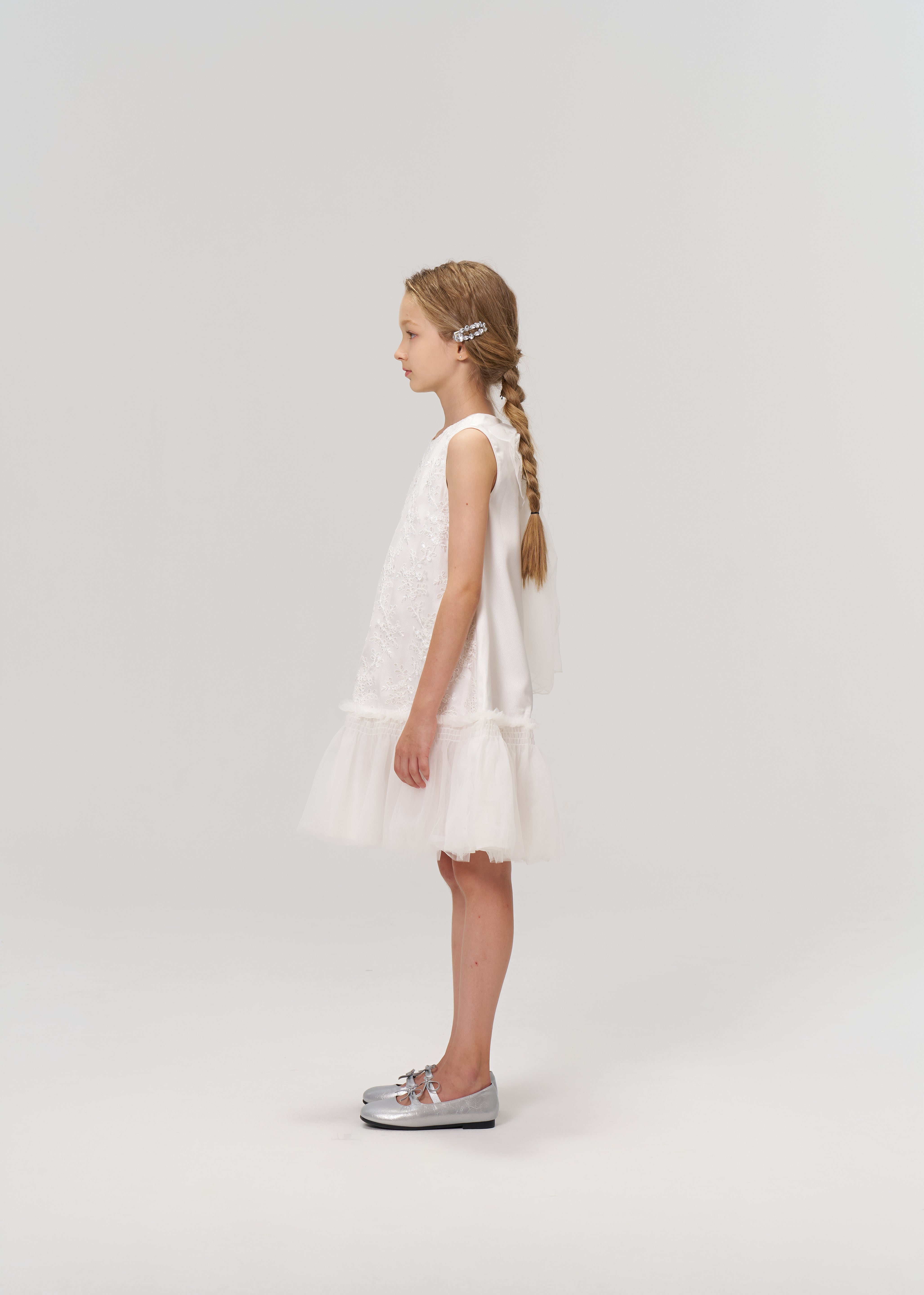 A young girl stands in profile against a light background, wearing the KIKI and JOJO Twinkle Whisper Dress—a white A-line style with an embroidered lace tulle skirt—paired with silver shoes.