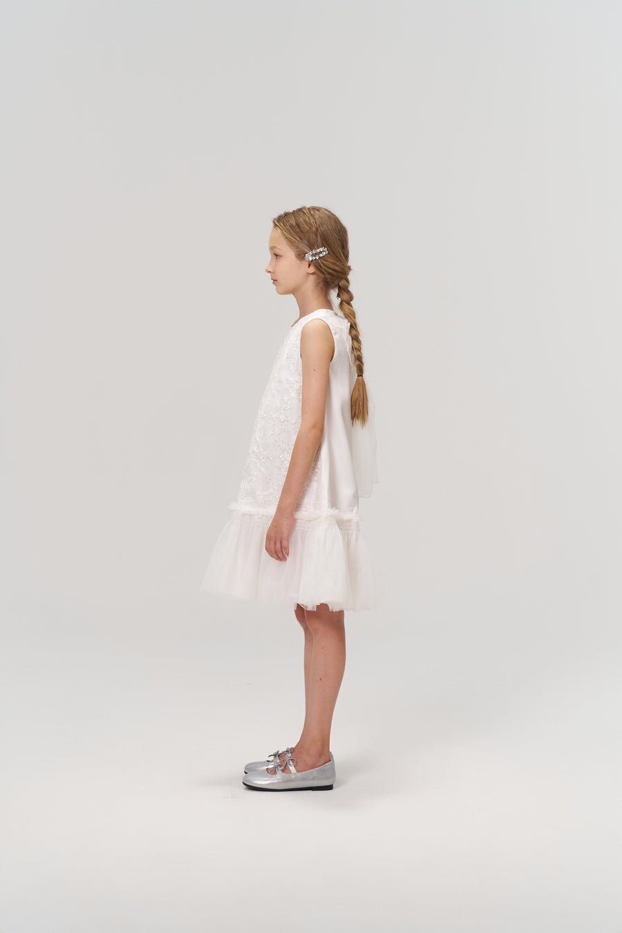 A young girl stands in profile against a light background, wearing the KIKI and JOJO Twinkle Whisper Dress—a white A-line style with an embroidered lace tulle skirt—paired with silver shoes.