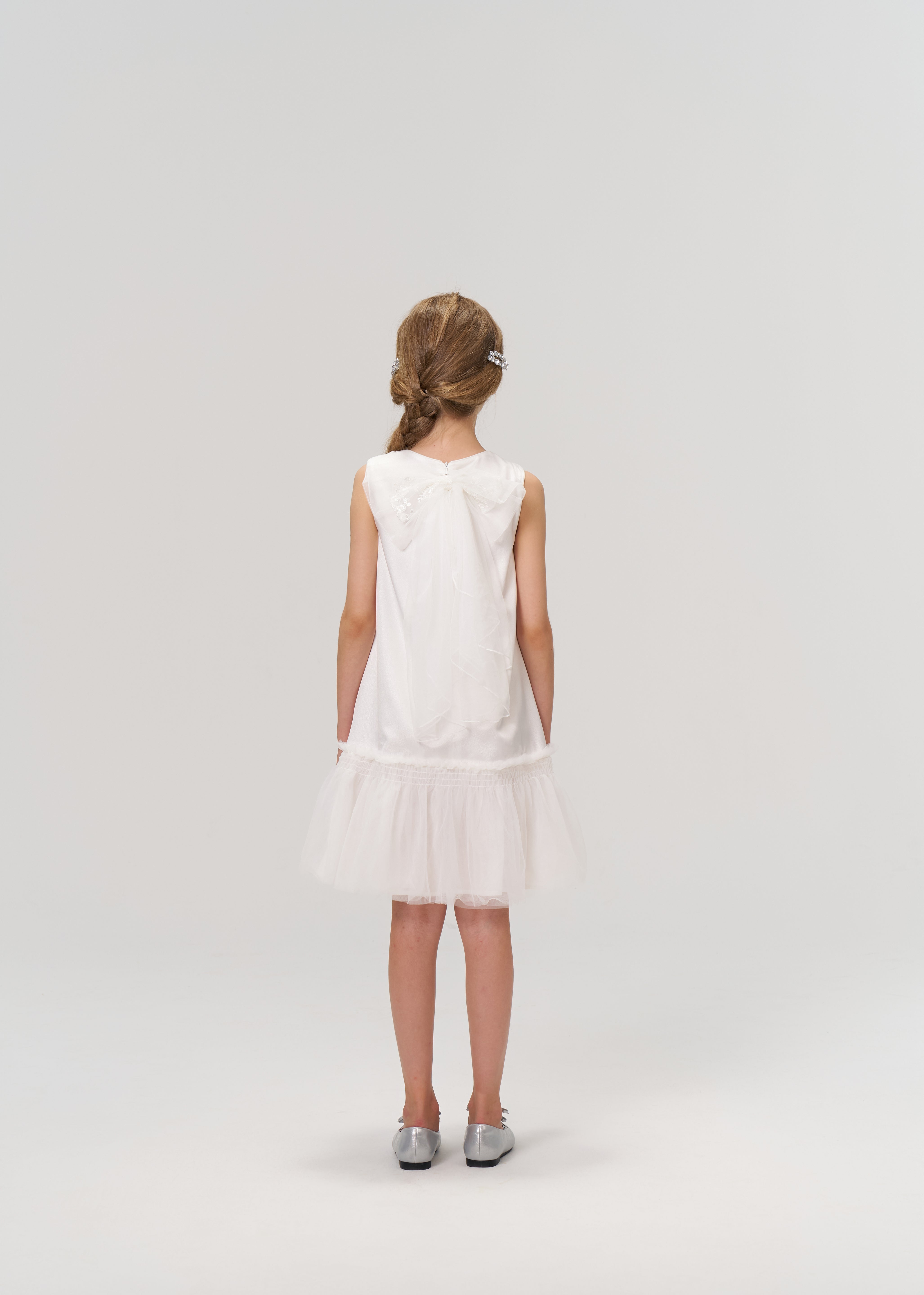 A young girl wearing the KIKI and JOJO Twinkle Whisper Dress stands facing away from the camera against a plain light background. She pairs the white A-line dress with embroidered lace details and silver shoes.