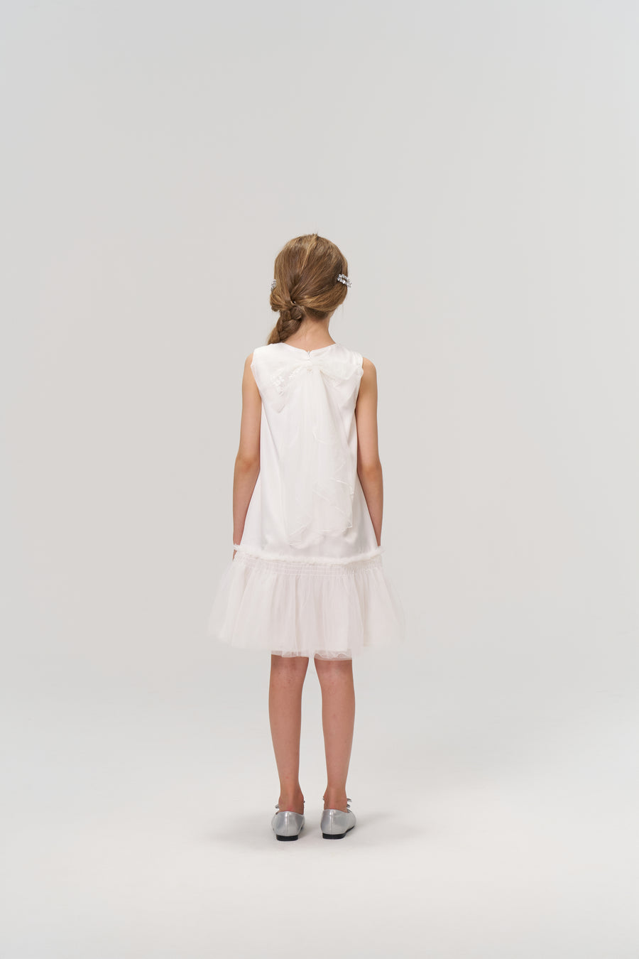A young girl wearing the KIKI and JOJO Twinkle Whisper Dress stands facing away from the camera against a plain light background. She pairs the white A-line dress with embroidered lace details and silver shoes.