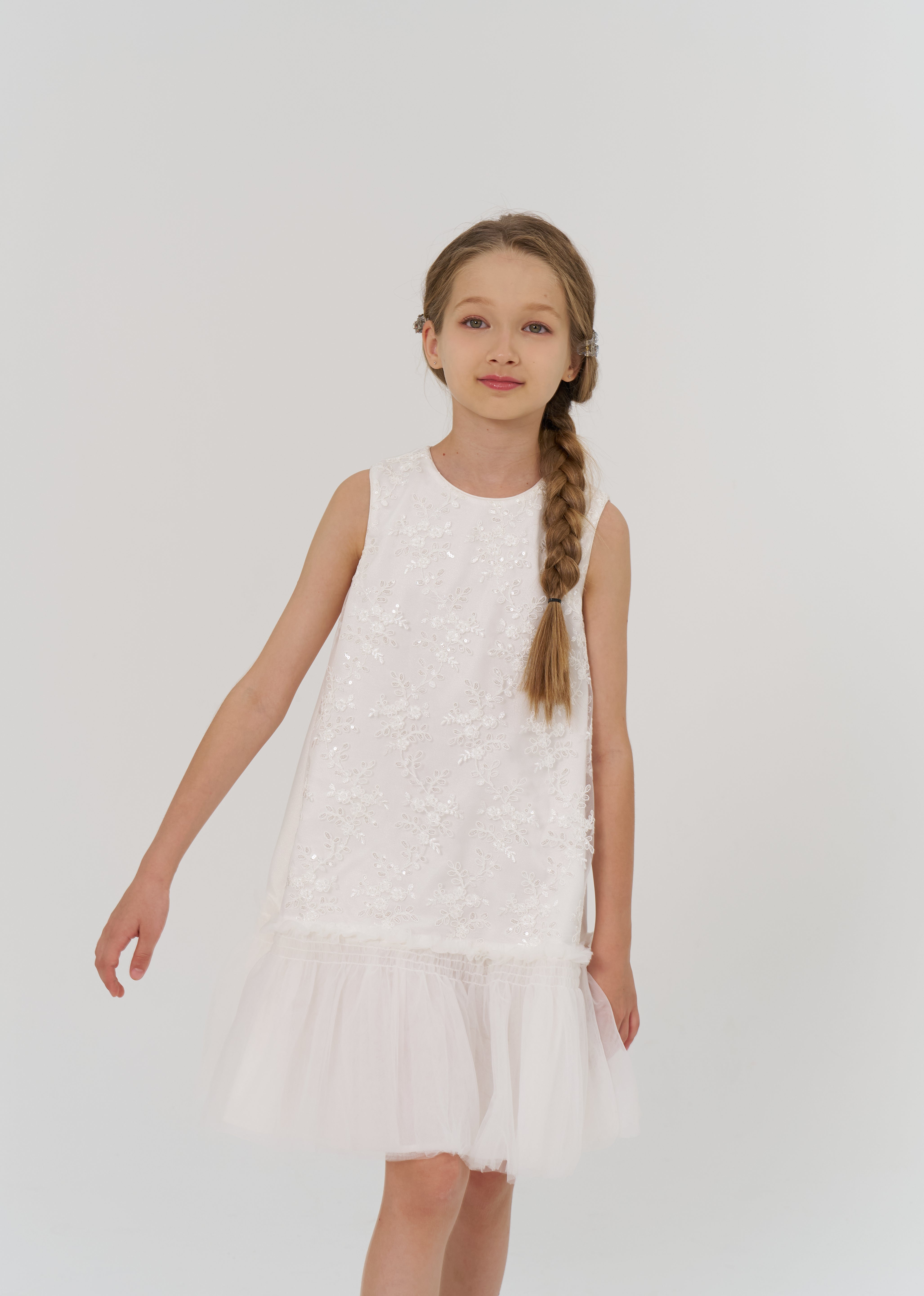 A young girl with a single braid wears the KIKI and JOJO Twinkle Whisper Dress, a sleeveless embroidered lace dress with tulle details, standing against a plain white background.