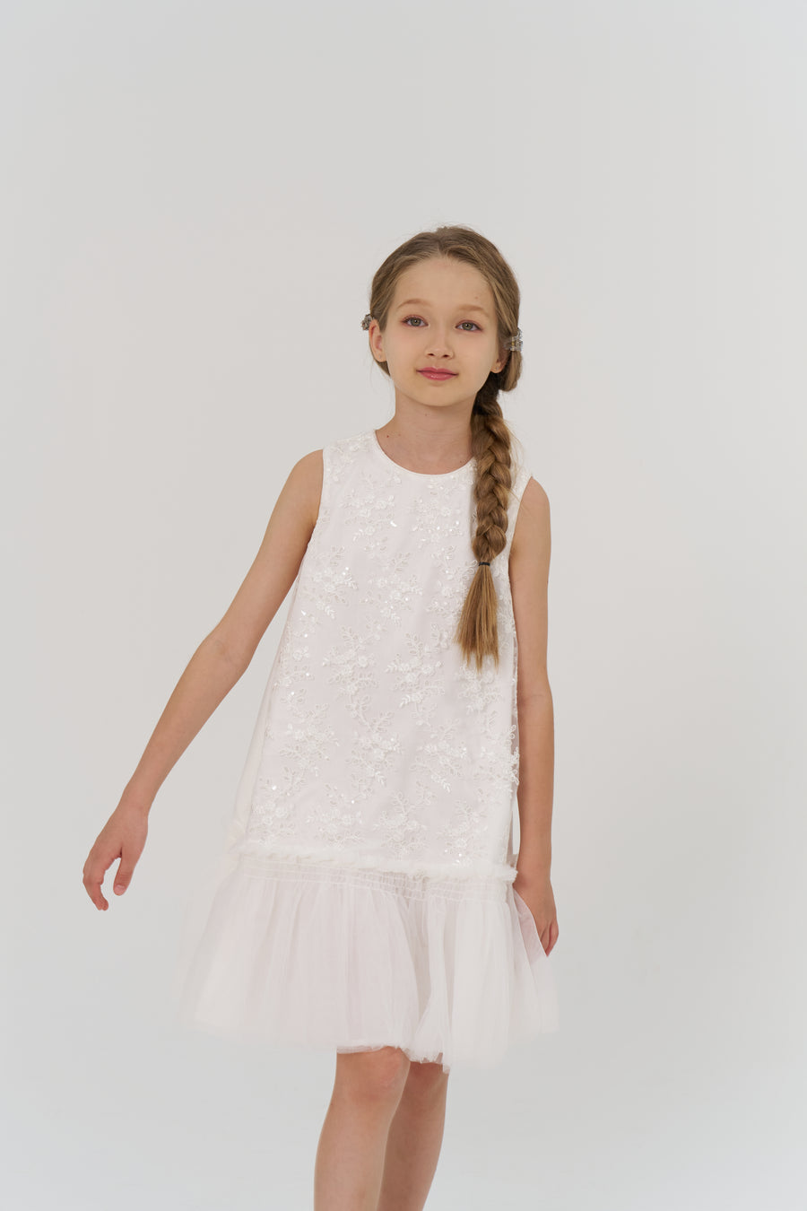 A young girl with a single braid wears the KIKI and JOJO Twinkle Whisper Dress, a sleeveless embroidered lace dress with tulle details, standing against a plain white background.