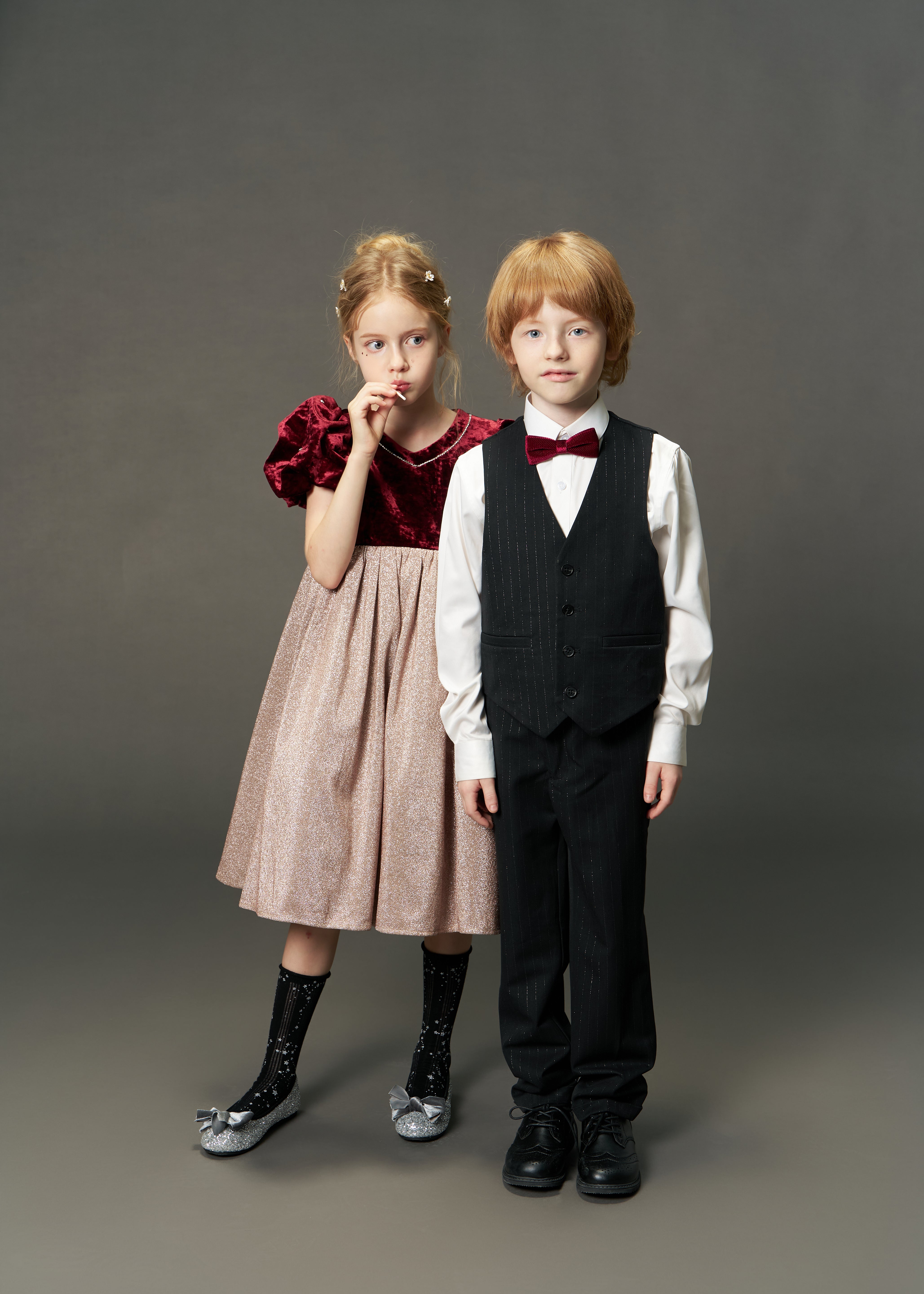 Two children stand side by side in formal attire; the girl wears the KIKI and JOJO Girls Velvet Puff Sleeve Dress with Rhinestone Trim in burgundy red, while the boy sports a suit with a bow tie against a plain background.