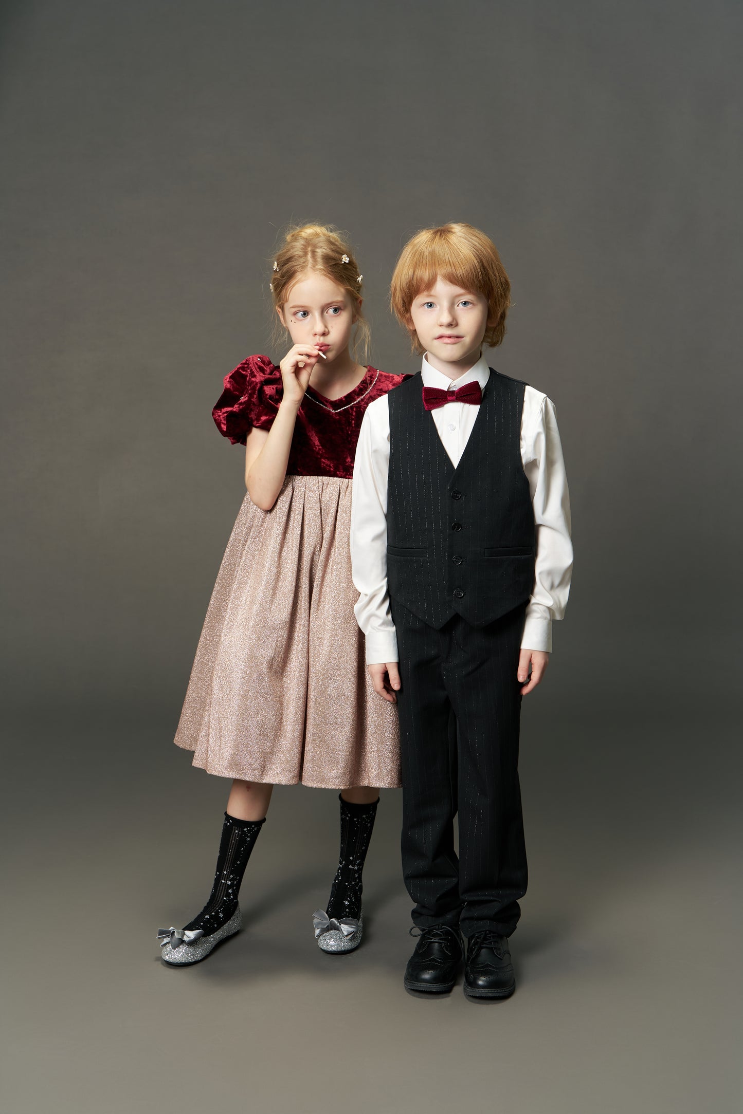 Two children stand side by side in formal attire; the girl wears the KIKI and JOJO Girls Velvet Puff Sleeve Dress with Rhinestone Trim in burgundy red, while the boy sports a suit with a bow tie against a plain background.