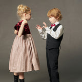 A young girl wearing the KIKI and JOJO Girls Velvet Puff Sleeve Dress with Rhinestone Trim in Burgundy Red holds a lollipop, standing across from a boy in a formal suit and bow tie against a plain background.