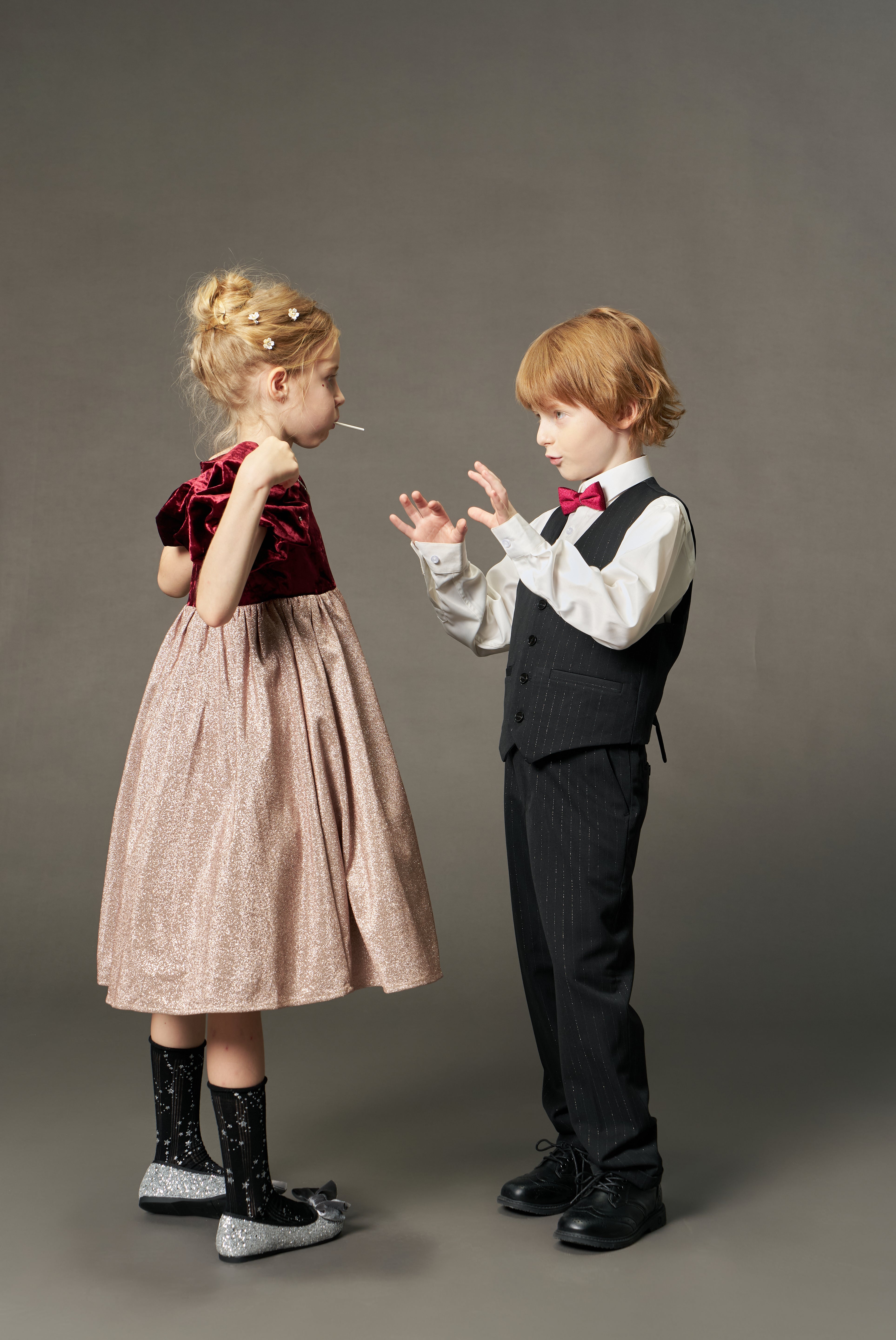 A young girl wearing the KIKI and JOJO Girls Velvet Puff Sleeve Dress with Rhinestone Trim in Burgundy Red holds a lollipop, standing across from a boy in a formal suit and bow tie against a plain background.