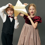 A young boy in a suit and a girl wearing the KIKI and JOJO Girls Velvet Puff Sleeve Dress with Rhinestone Trim in Burgundy Red hold a gold glittery star together against a plain gray background.