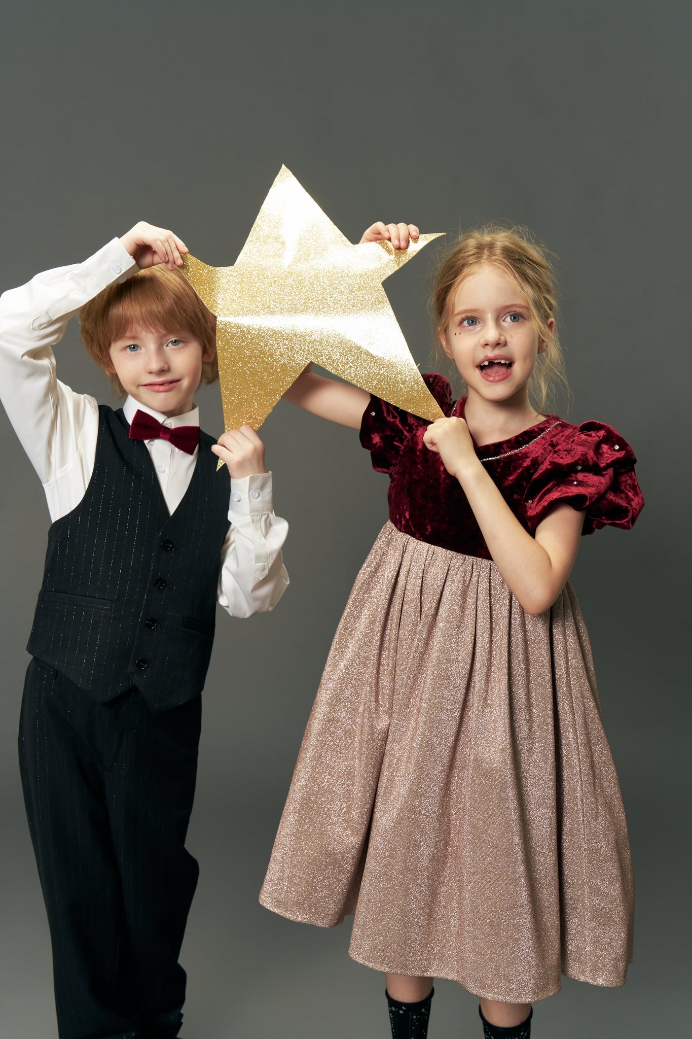 A young boy in a suit and a girl wearing the KIKI and JOJO Girls Velvet Puff Sleeve Dress with Rhinestone Trim in Burgundy Red hold a gold glittery star together against a plain gray background.