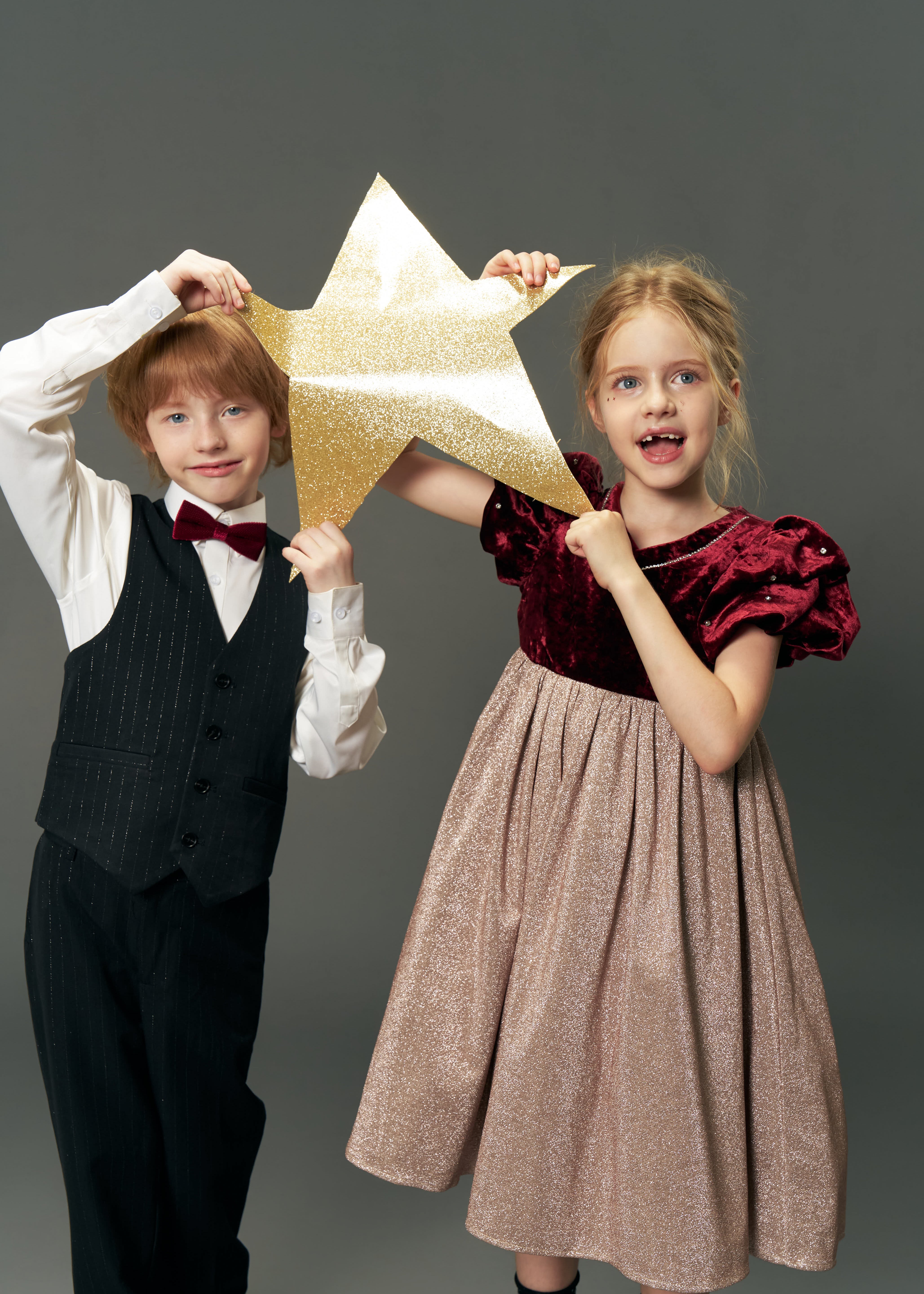 A young boy in a suit and a girl wearing the KIKI and JOJO Girls Velvet Puff Sleeve Dress with Rhinestone Trim in Burgundy Red hold a gold glittery star together against a plain gray background.
