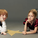 A boy and a girl lie on the floor with a large gold star between them; the girl wears the KIKI and JOJO Girls Velvet Puff Sleeve Dress with Rhinestone Trim in Burgundy Red, both looking at the camera against a plain gray background.