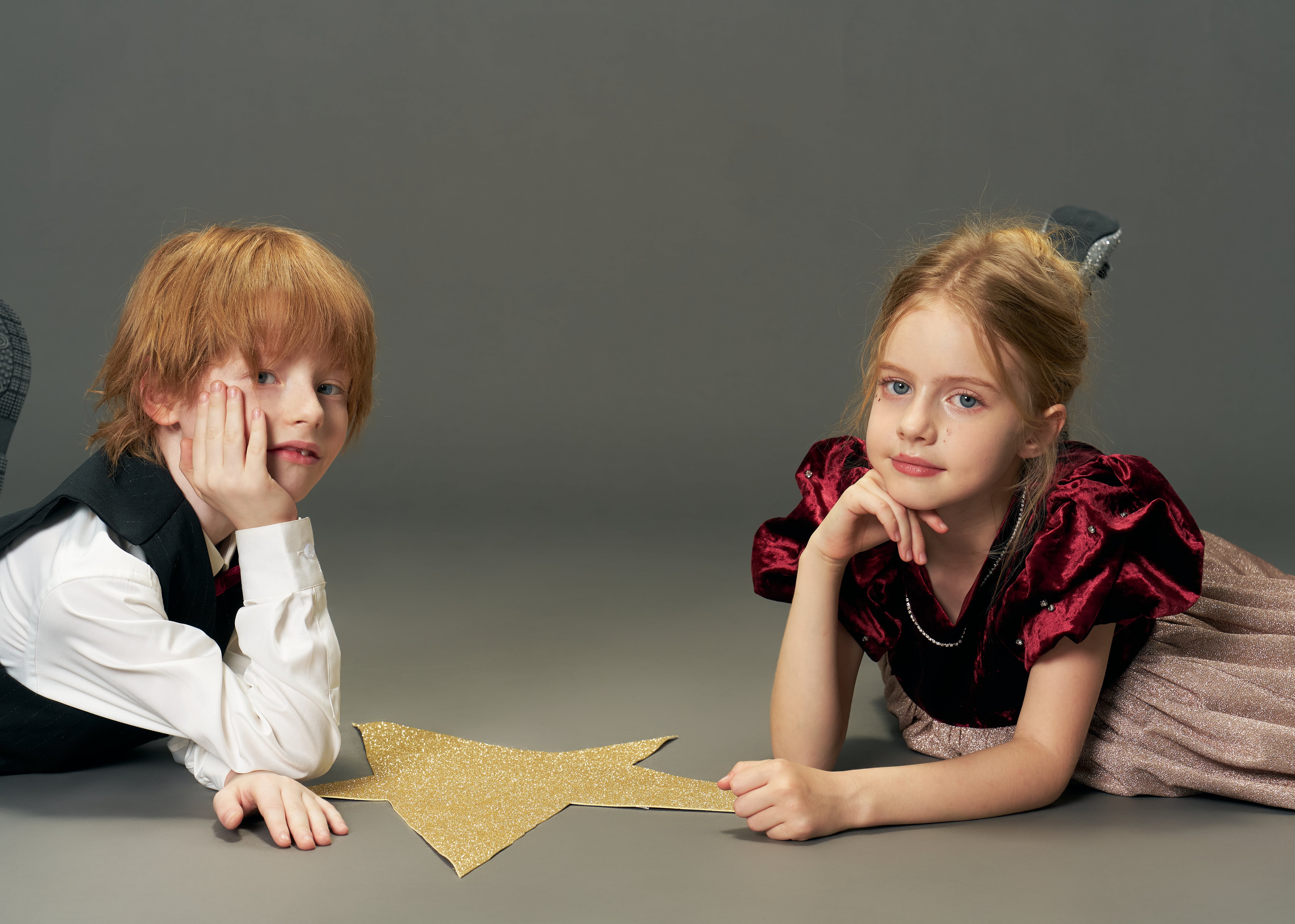A boy and a girl lie on the floor with a large gold star between them; the girl wears the KIKI and JOJO Girls Velvet Puff Sleeve Dress with Rhinestone Trim in Burgundy Red, both looking at the camera against a plain gray background.